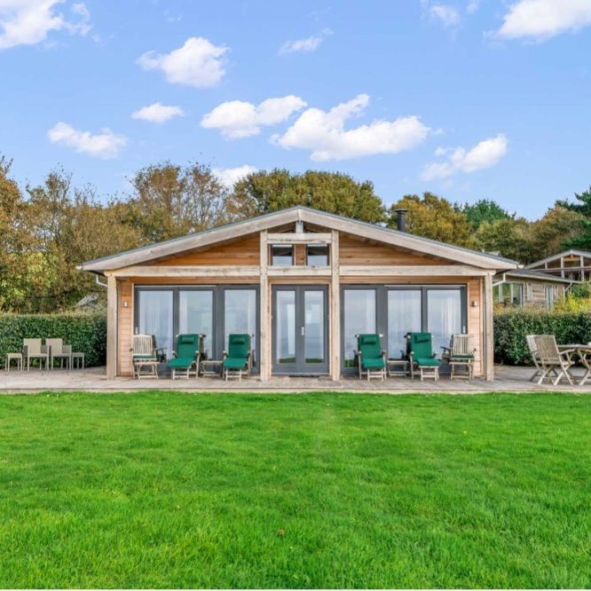 Modern wooden cabin with large windows and patio furniture, surrounded by greenery and trees under a blue sky.