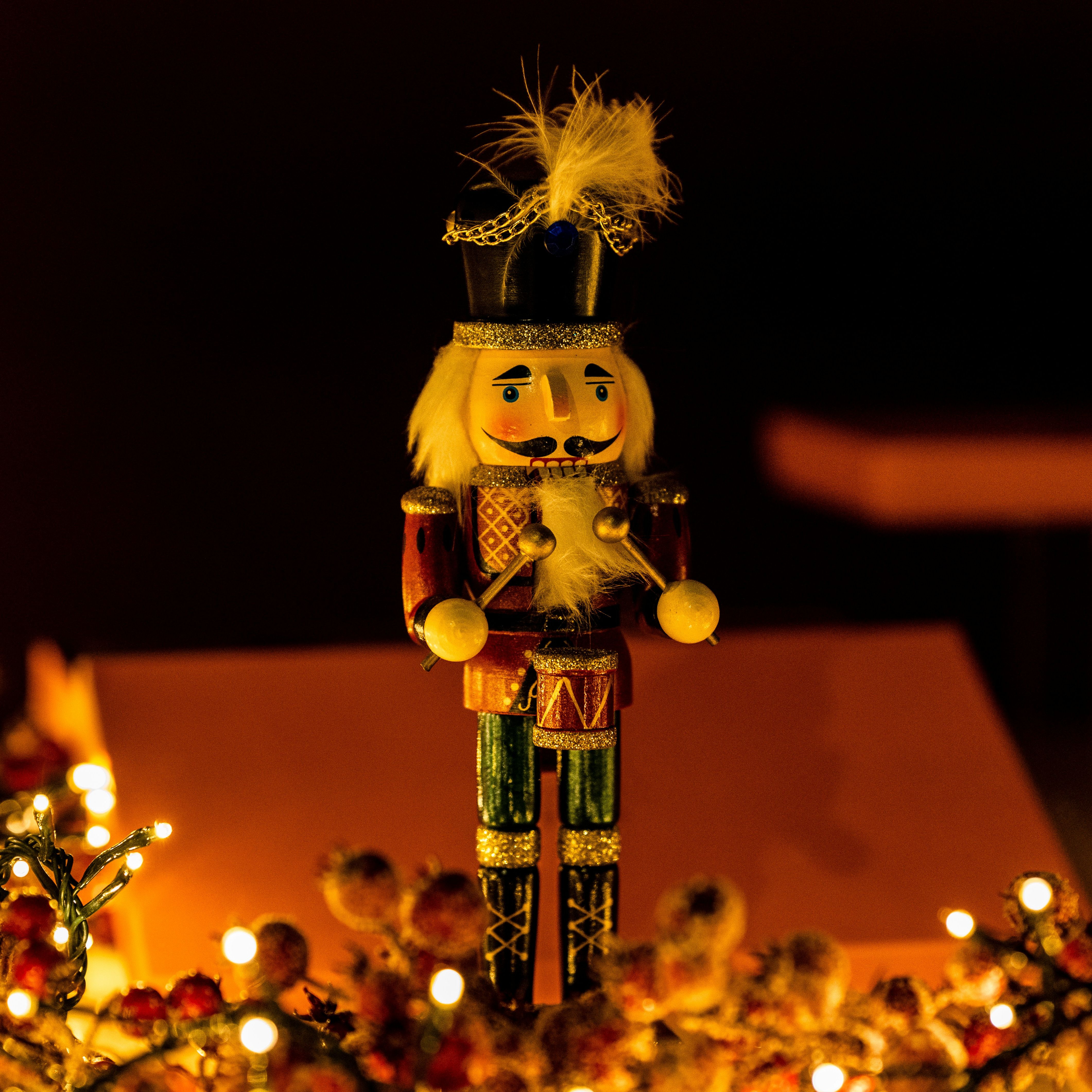 A wooden nutcracker figurine with a black hat and drum, surrounded by festive lights and decorations.