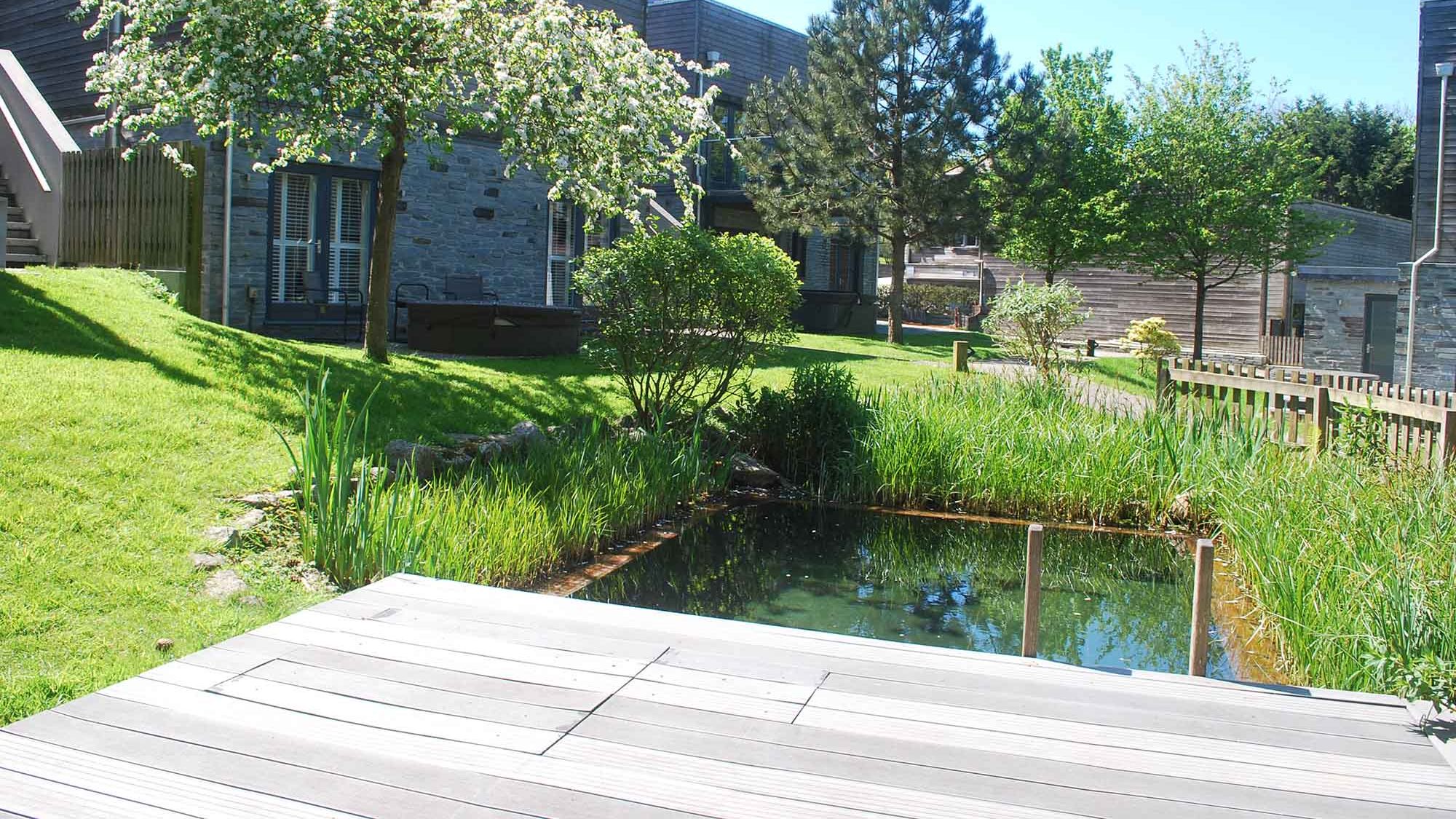 Garden with pond, trees, green grass, and modern stone and wood buildings in the background.
