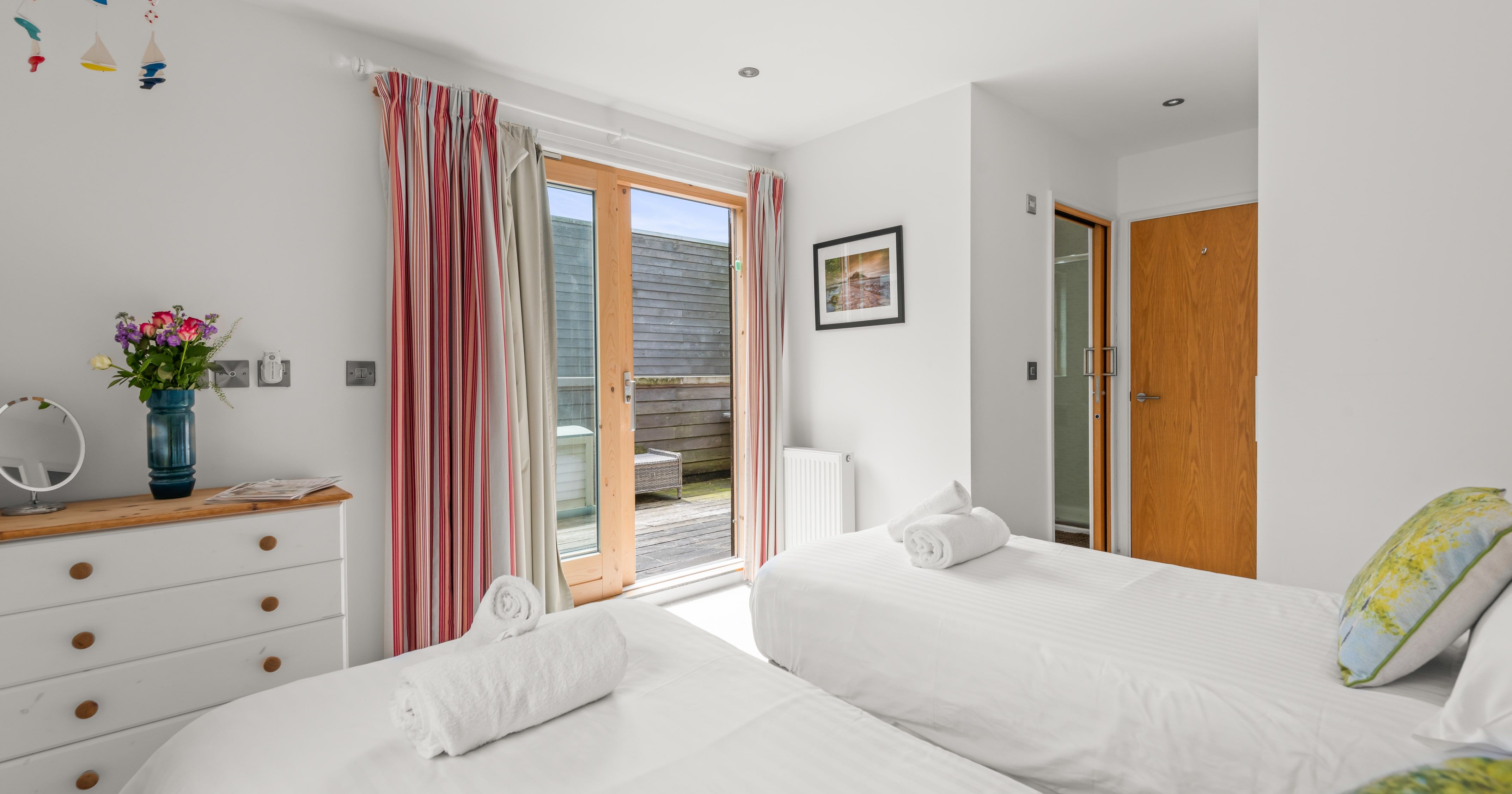 Bright modern bedroom with two single beds, white bedding, striped curtains, a dresser with a mirror and flowers, wooden doors, and access to a small outdoor patio.