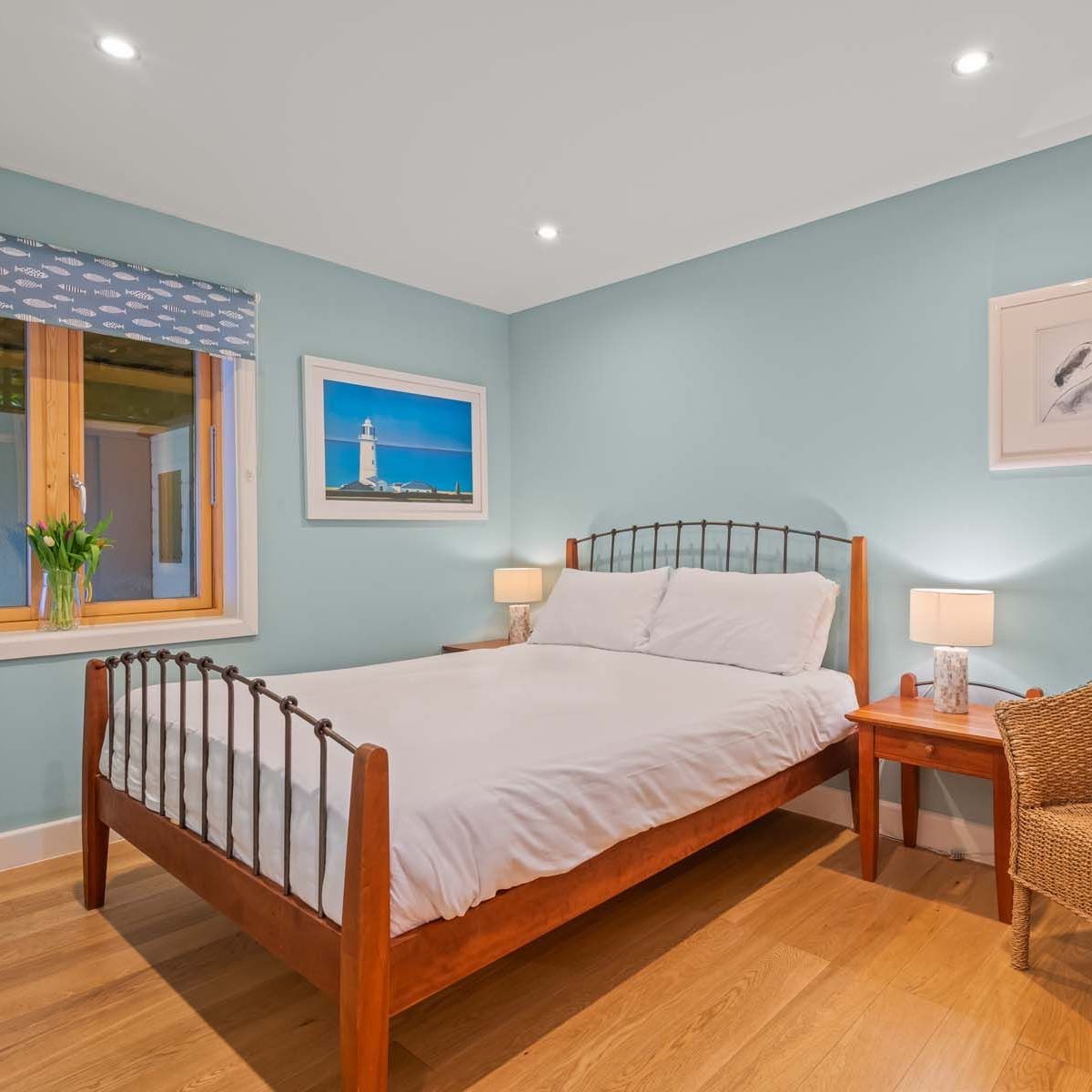 Cozy bedroom with a wooden bed frame, wicker chair, light blue walls, and framed artwork.