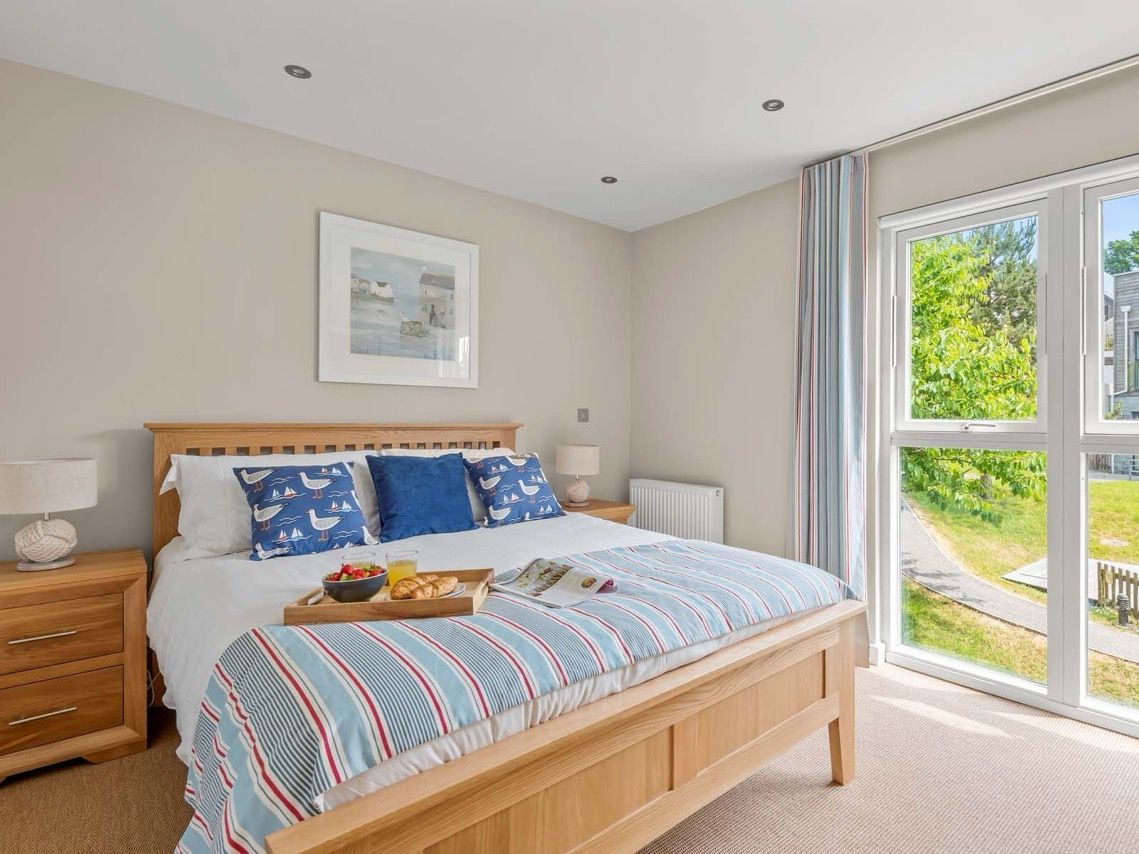 Bright, cozy bedroom with wooden bed, blue and white bedding, large windows, and a breakfast tray on the bed.