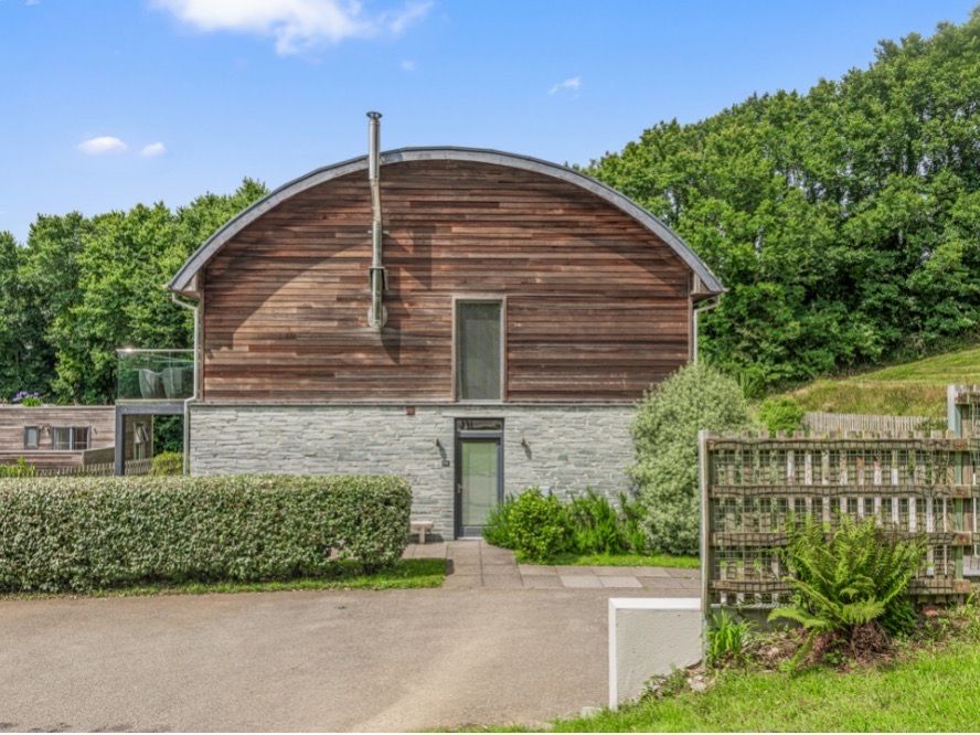 Modern house with curved wooden roof and stone facade, surrounded by greenery.