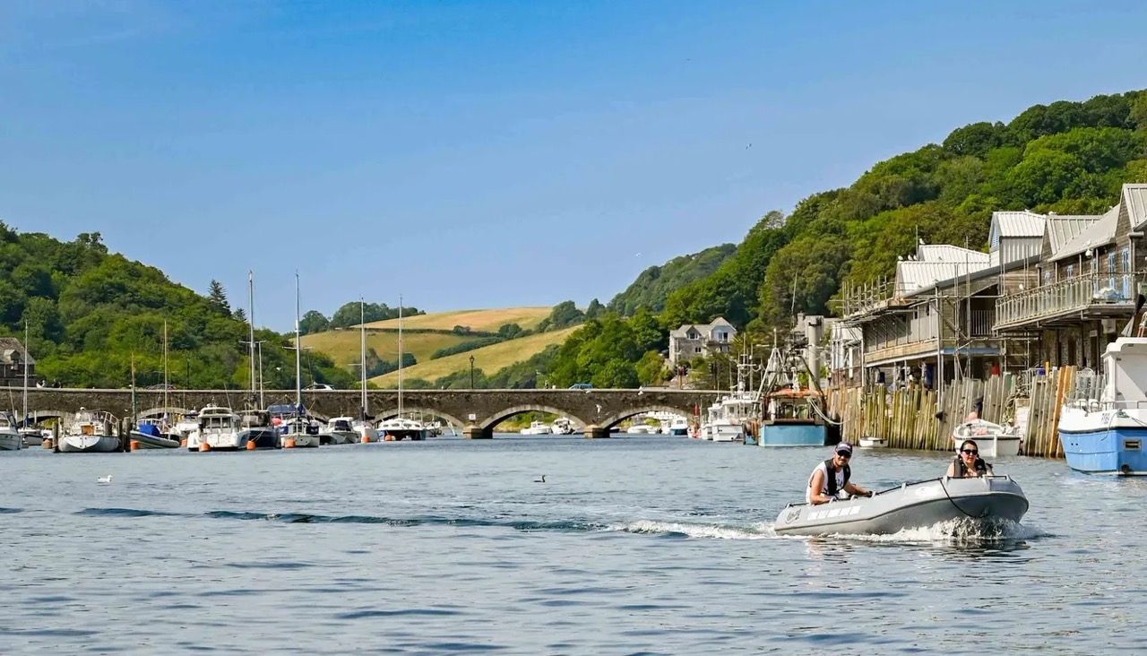 People riding a small motorboat on a scenic river, with boats docked along the shoreline, a stone bridge, and green hills in the background.