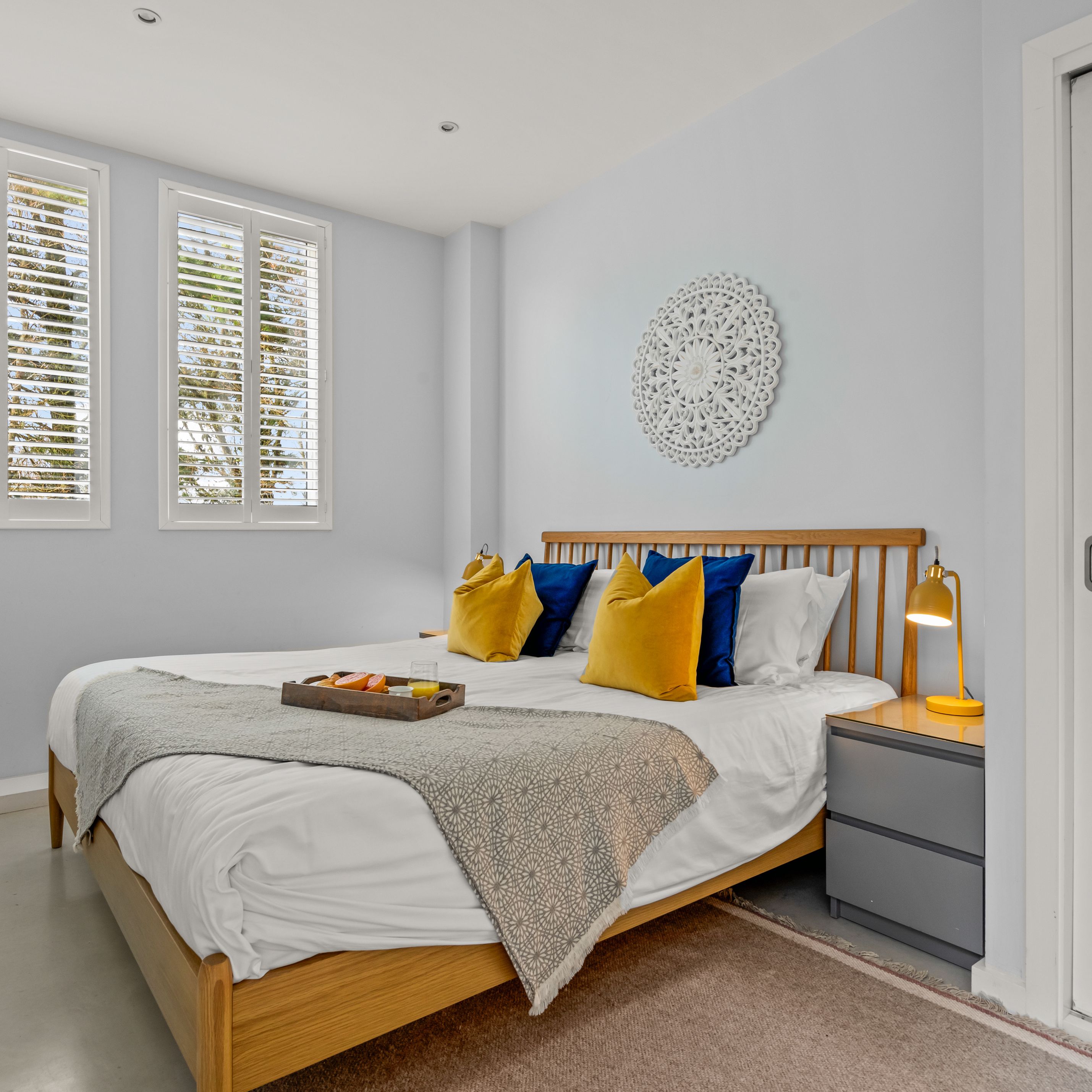 Bright minimalist bedroom with wooden bed, yellow and blue cushions, and white shutters