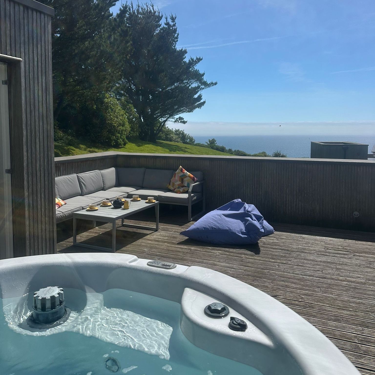 Hot tub on a wooden terrace with outdoor sofa, beanbag and sea view under a blue sky