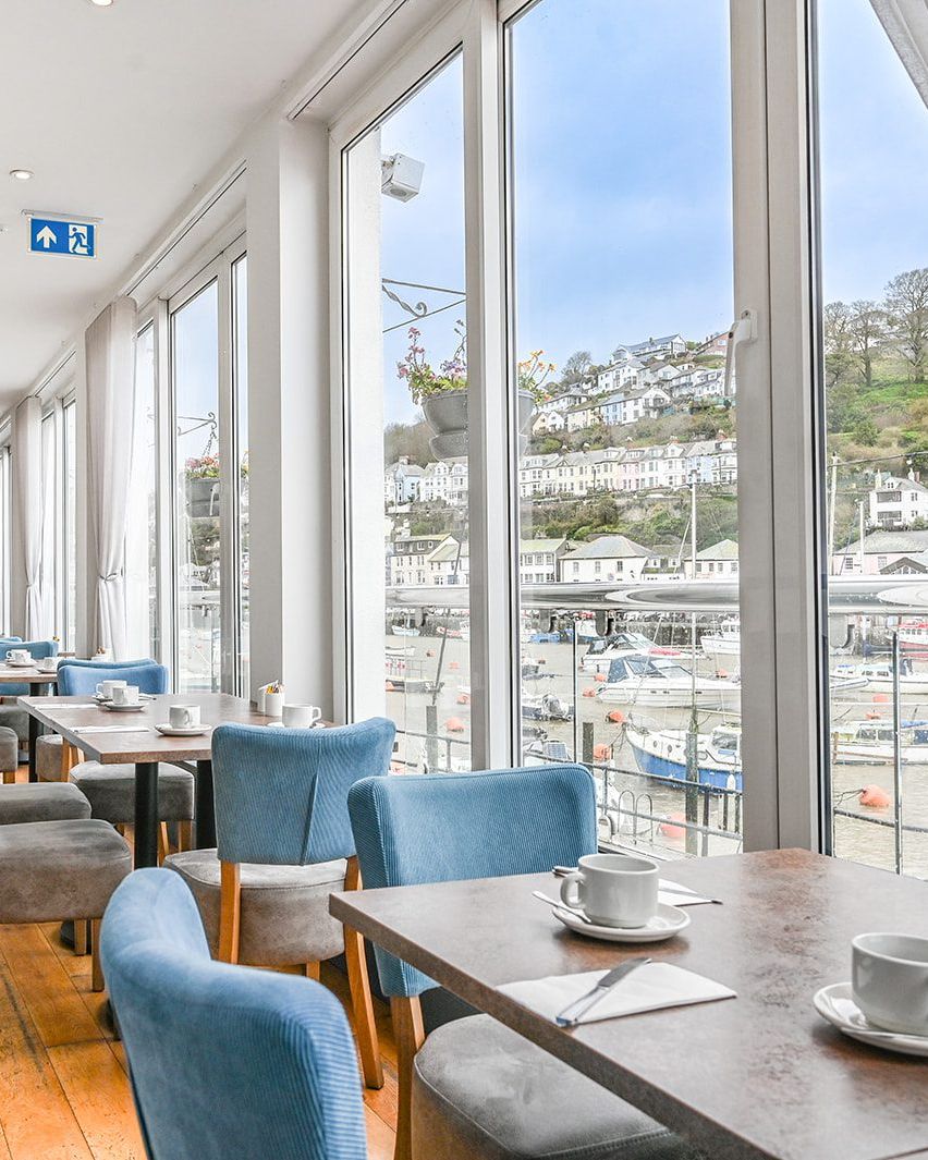 Bright waterfront café dining area with blue chairs and tables set with teacups beside large windows overlooking a harbour.