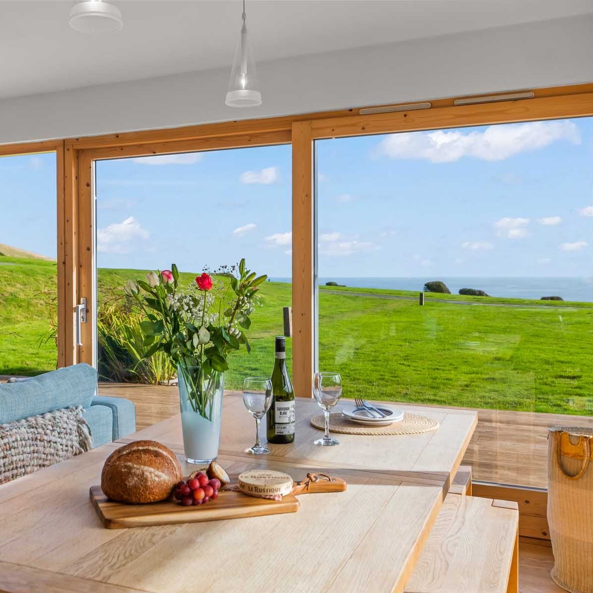 Light-filled dining area with wooden-framed windows overlooking green coastal fields and the sea