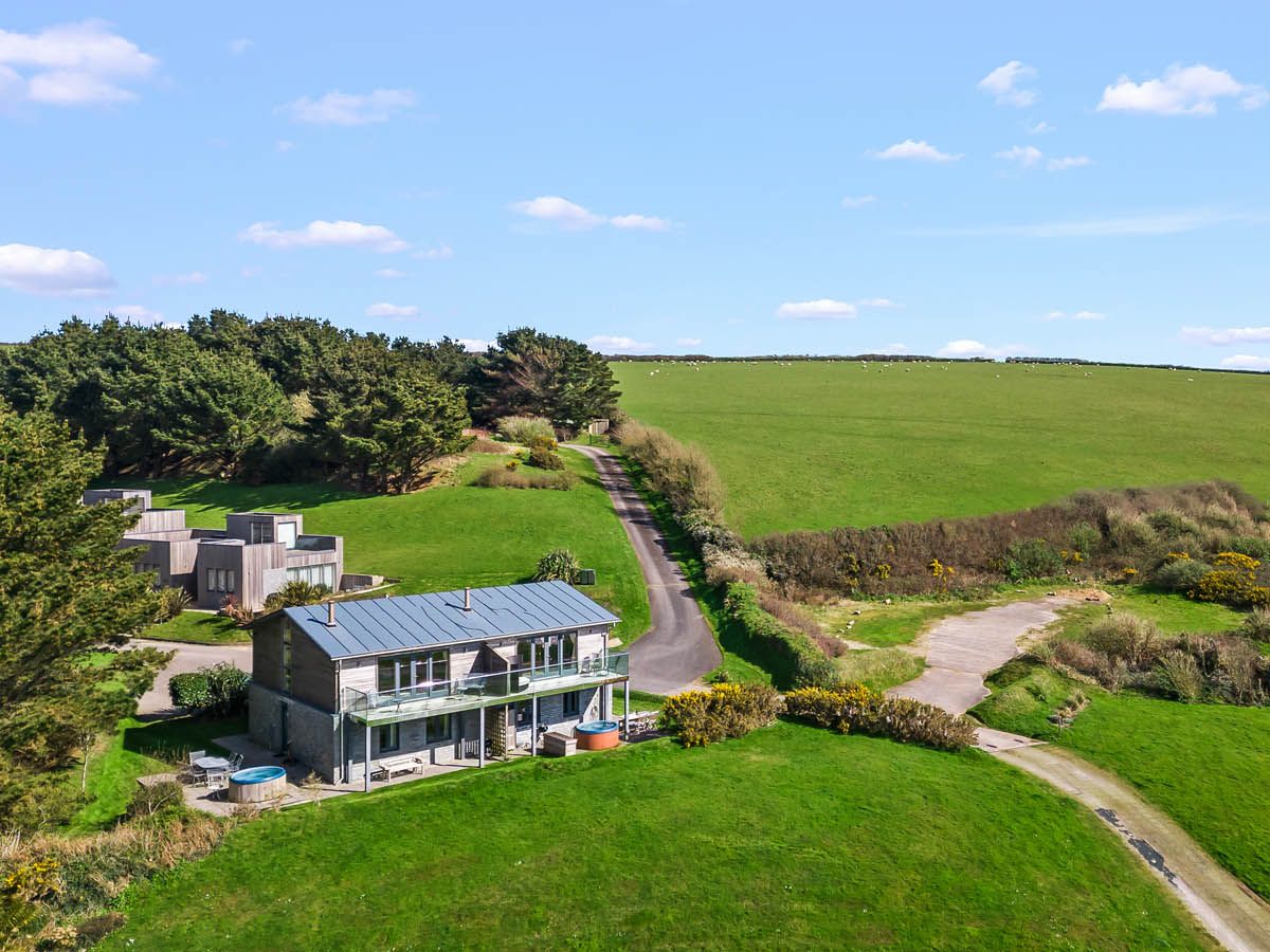 Aerial view of a modern countryside house with a metal roof beside a winding lane and green fields under a blue sky