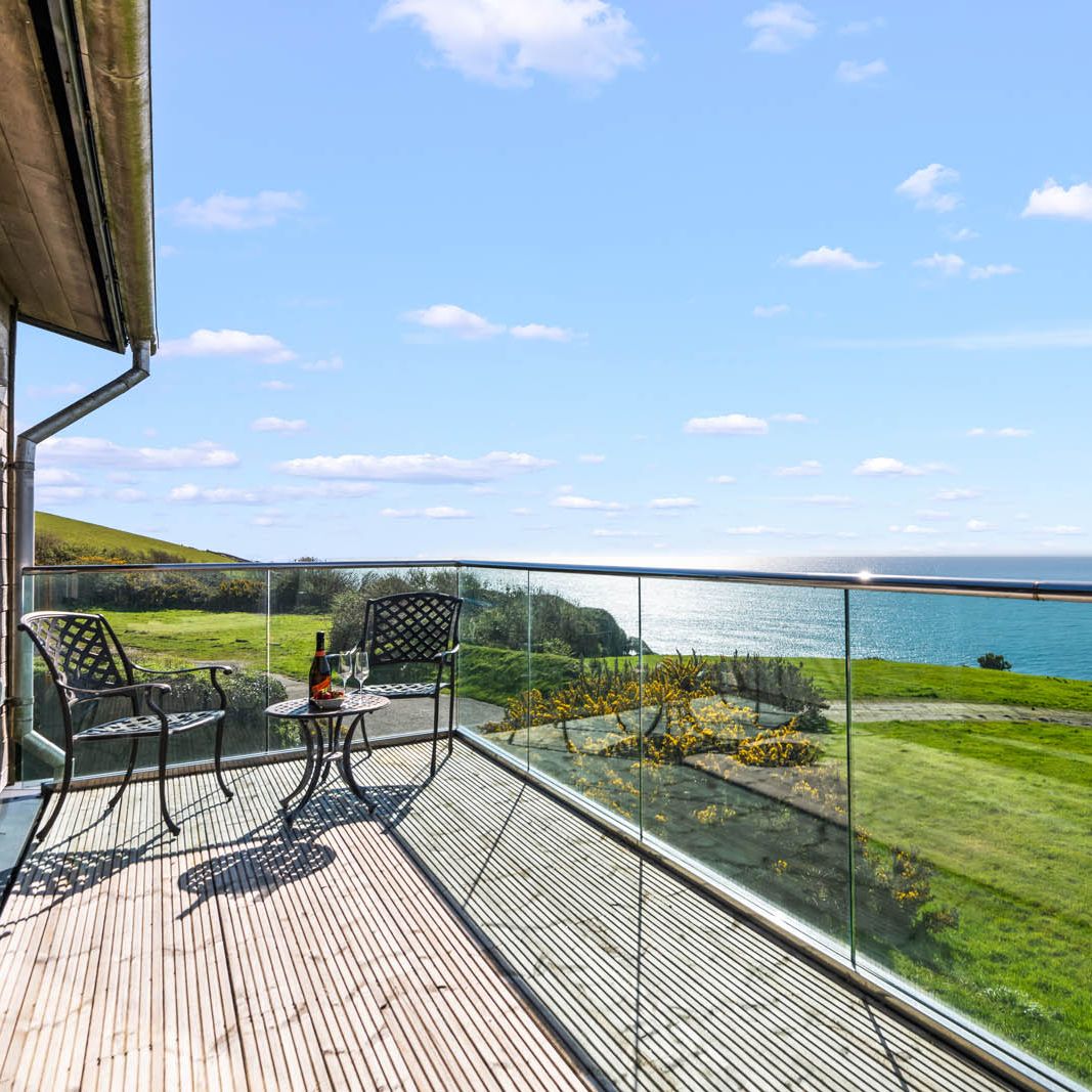 Balcony terrace with two chairs and a small table overlooking green hills and the sea under a blue sky