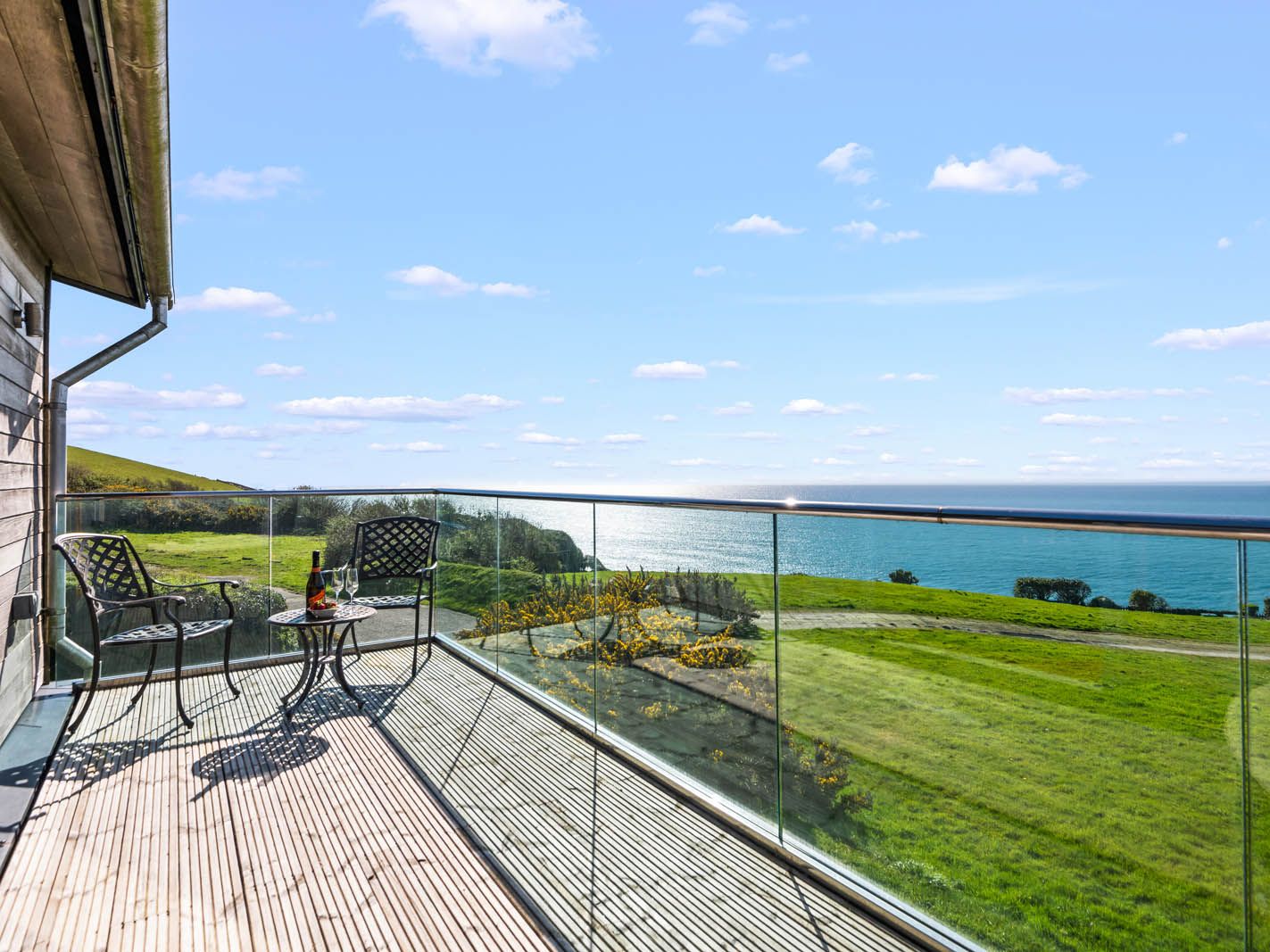 Balcony terrace with two chairs and a small table overlooking green hills and the sea under a blue sky