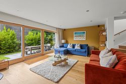 Modern living room with large glass doors opening to a garden patio, featuring a blue sofa, red sofa, and round glass coffee table.