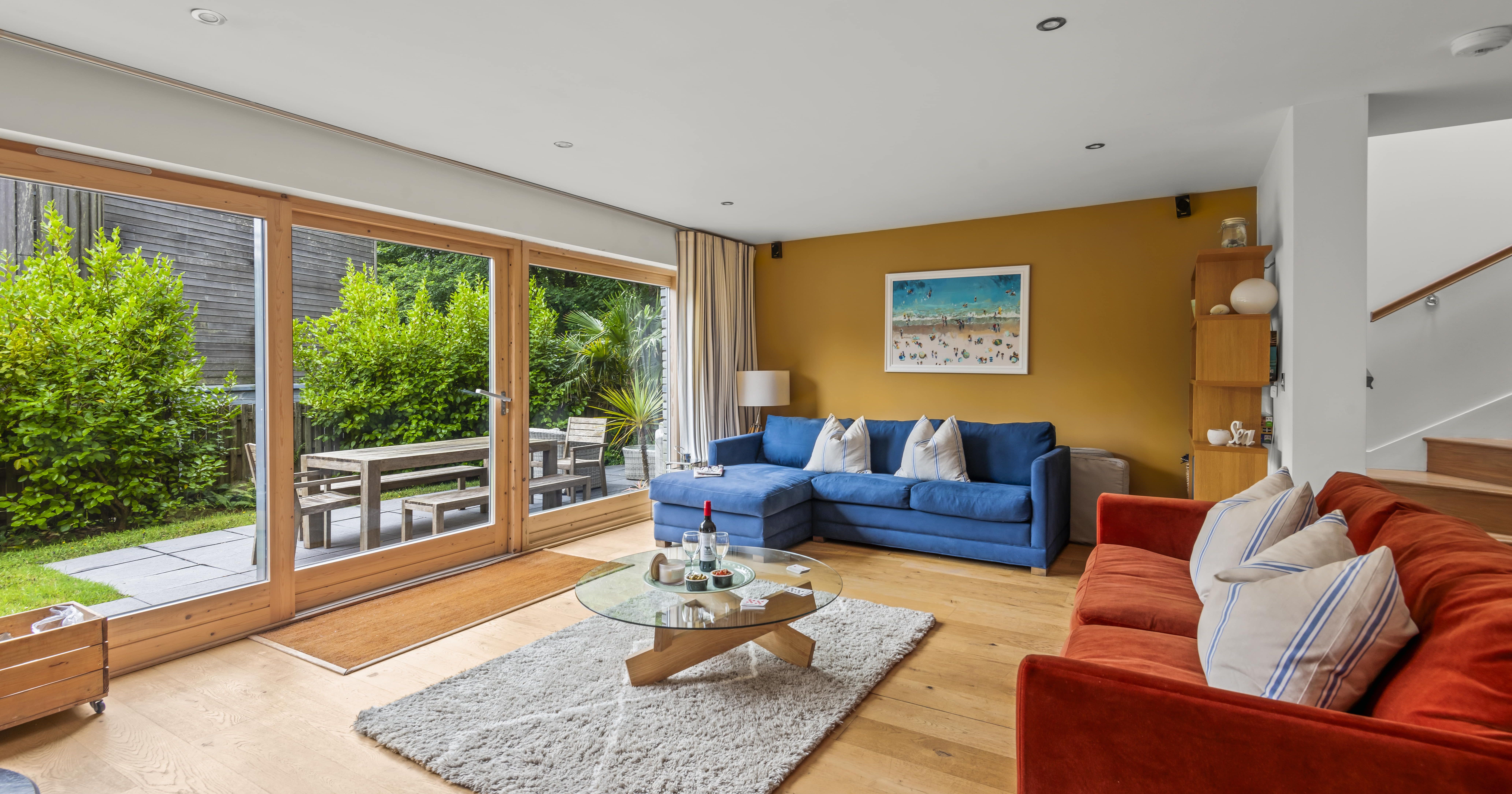 Modern living room with large glass doors opening to a garden patio, featuring a blue sofa, red sofa, and round glass coffee table.