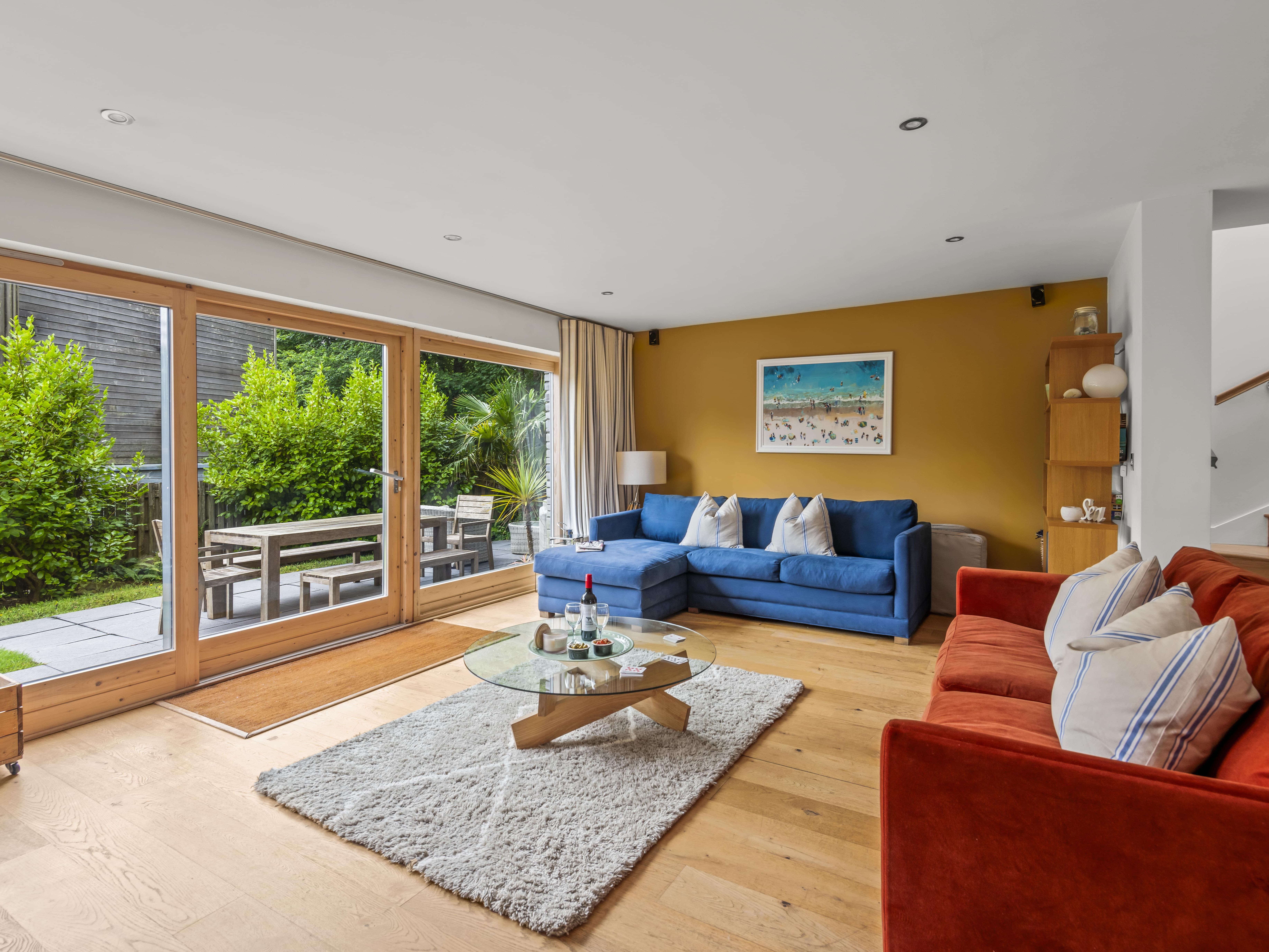 Modern living room with large glass doors opening to a garden patio, featuring a blue sofa, red sofa, and round glass coffee table.