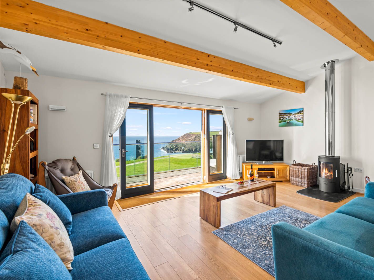 Bright coastal living room with blue sofas, wood-burning stove and glass doors opening to a sea-view terrace