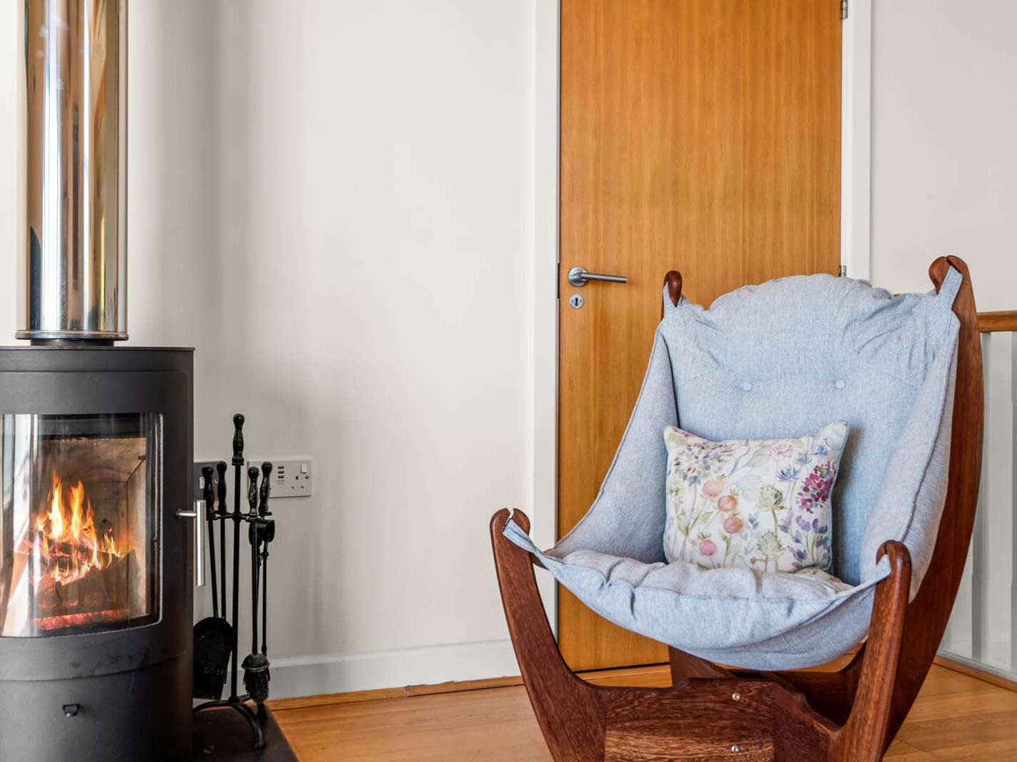 Wood-burning stove with a lit fire beside a cushioned wooden armchair and a closed wooden door