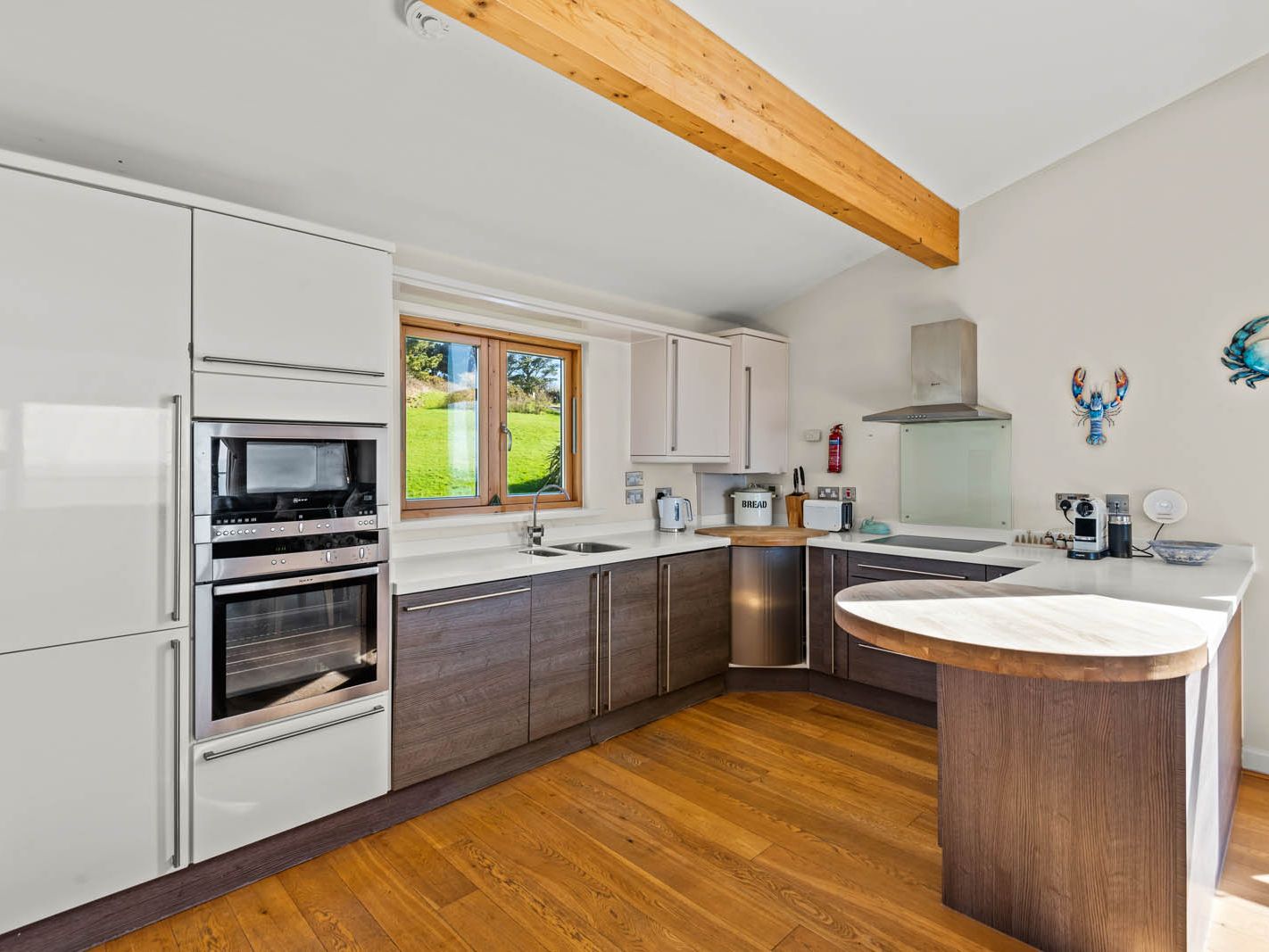 Modern kitchen with white and dark wood cabinets, built-in oven and hob, wooden floor and breakfast bar, with a window overlooking a green hillside