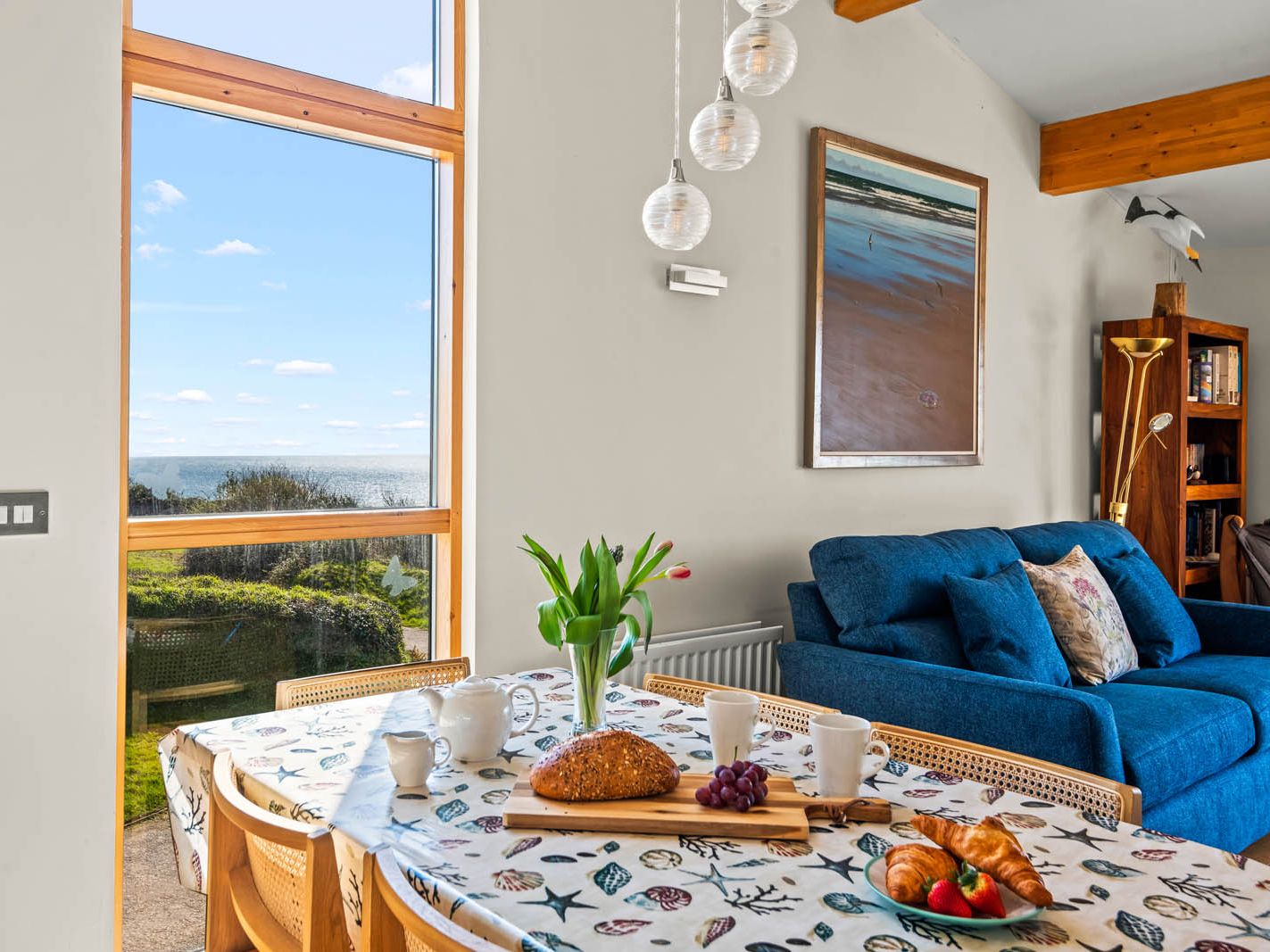 Bright coastal dining area with a table set for breakfast beside a blue sofa and large window overlooking the sea
