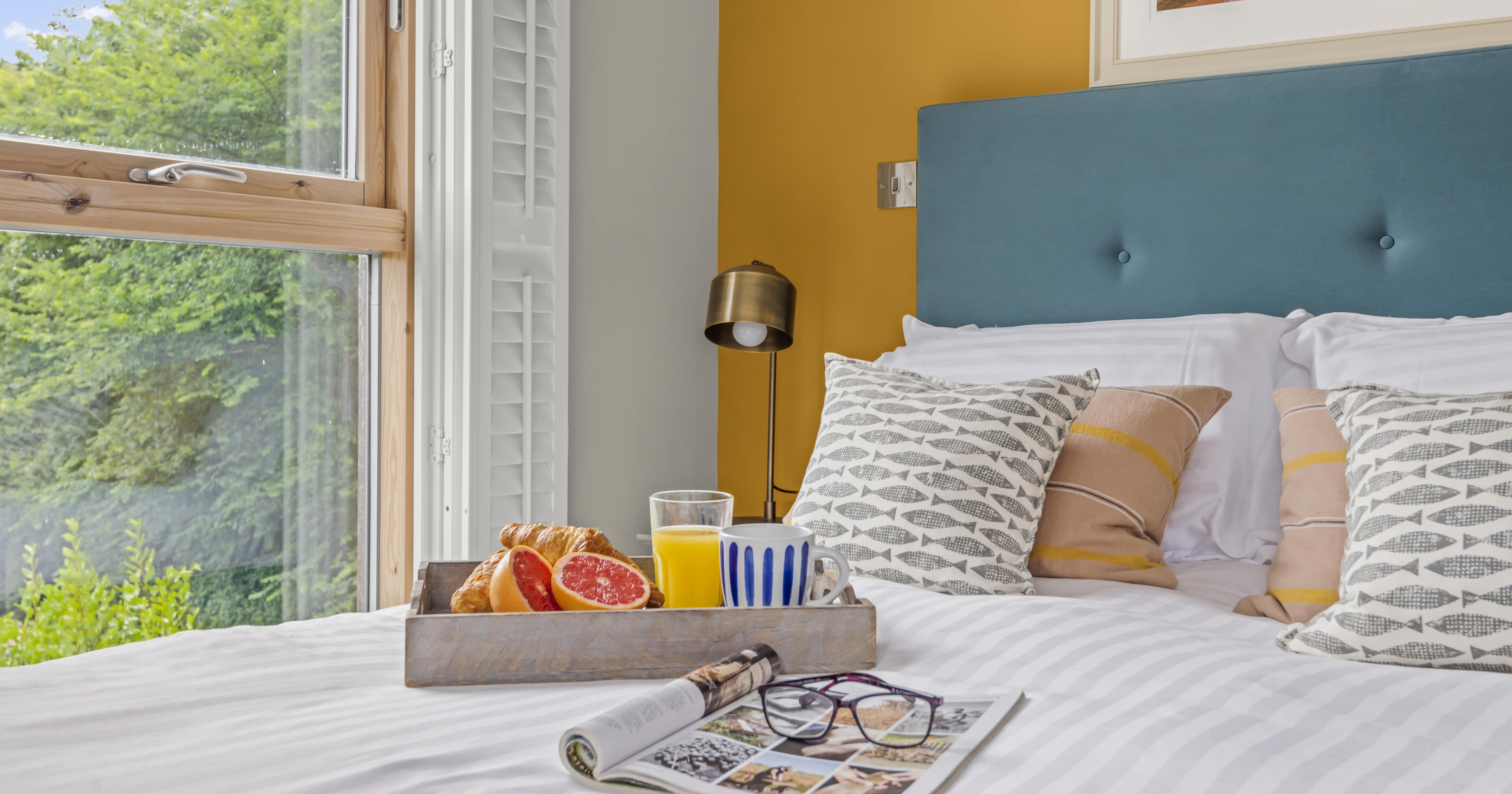 Breakfast tray with croissant, grapefruit, orange juice and coffee on a bed with decorative pillows, beside a window with a garden view