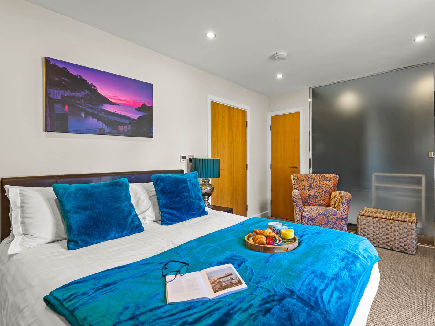 Modern hotel bedroom with turquoise bedding, breakfast tray on the bed, and en-suite sink area behind a frosted glass panel
