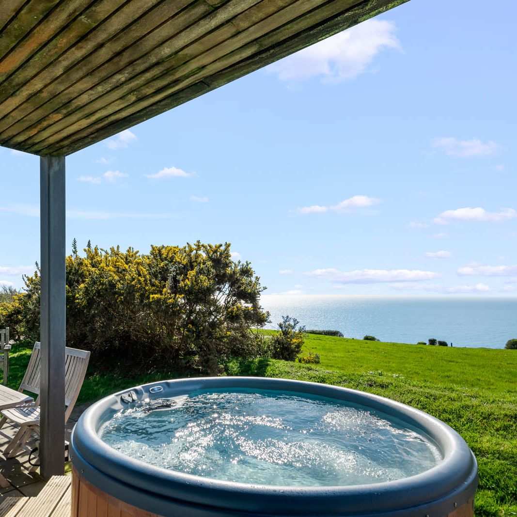 Hot tub on a patio under a pergola overlooking green fields and the sea on a sunny day