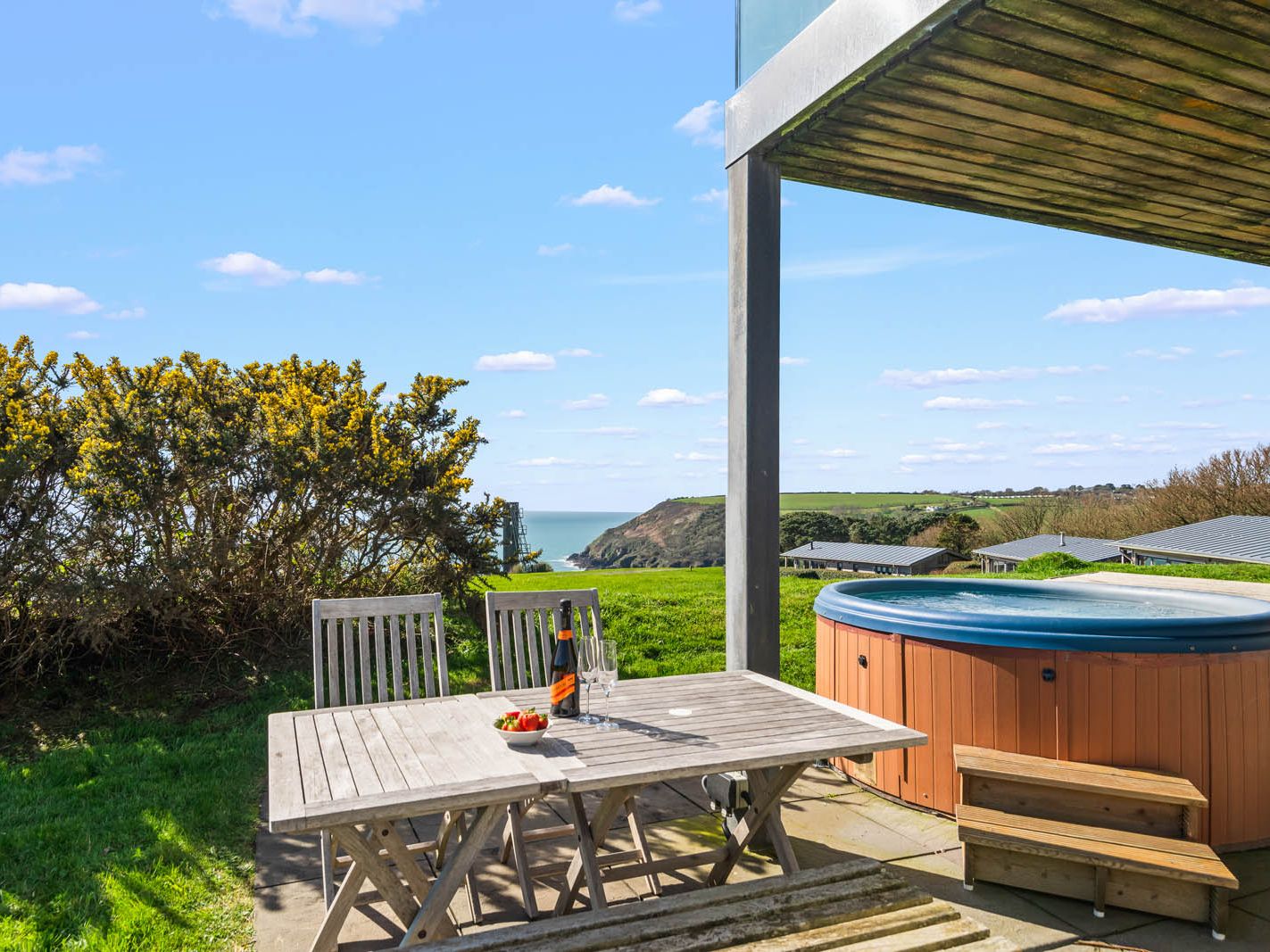 Patio table set beside a hot tub with coastal sea views and green fields under a blue sky