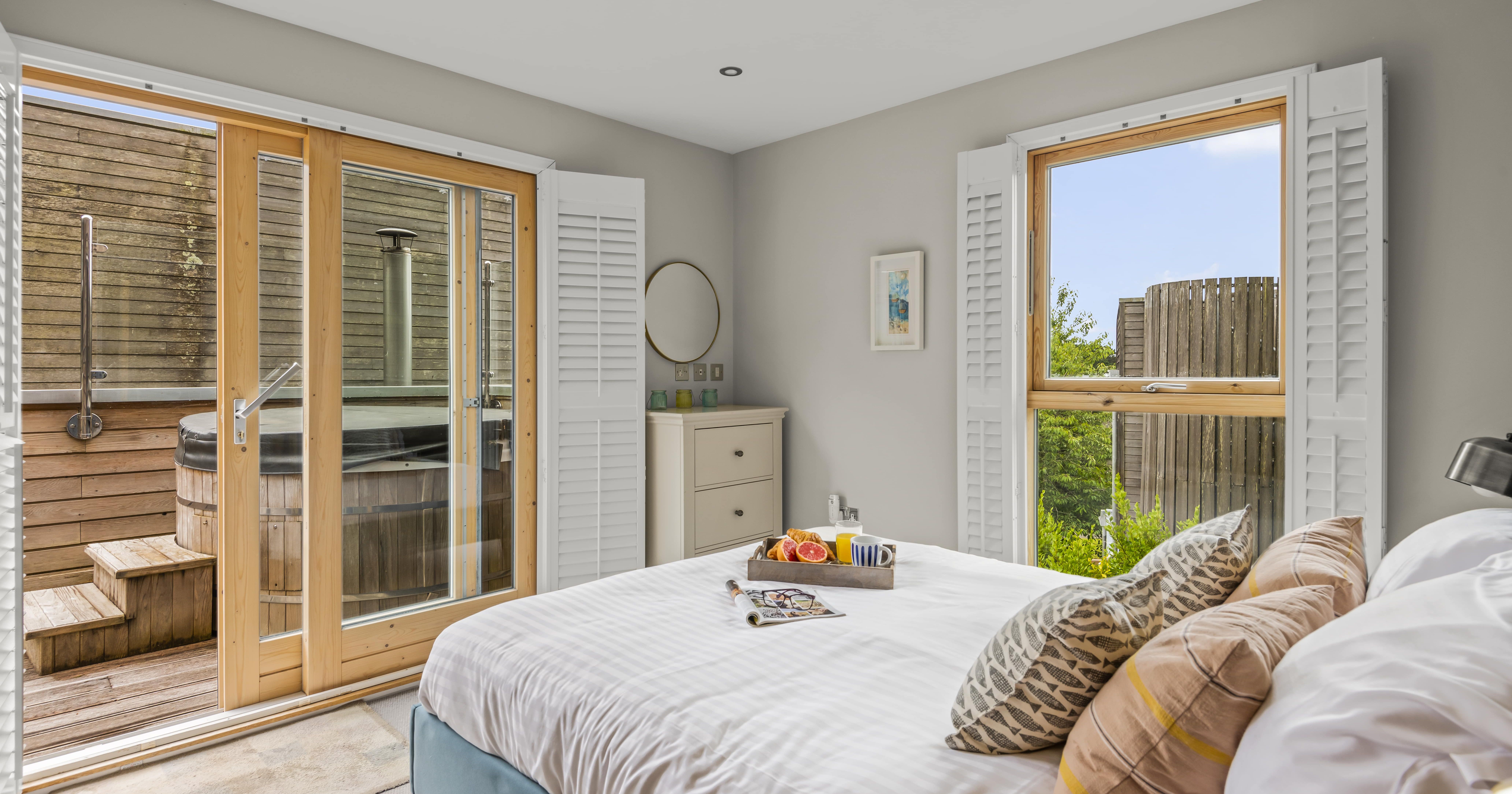 Bright bedroom with white bedding, wooden doors leading to outdoor wooden hot tub, and window shutters.
