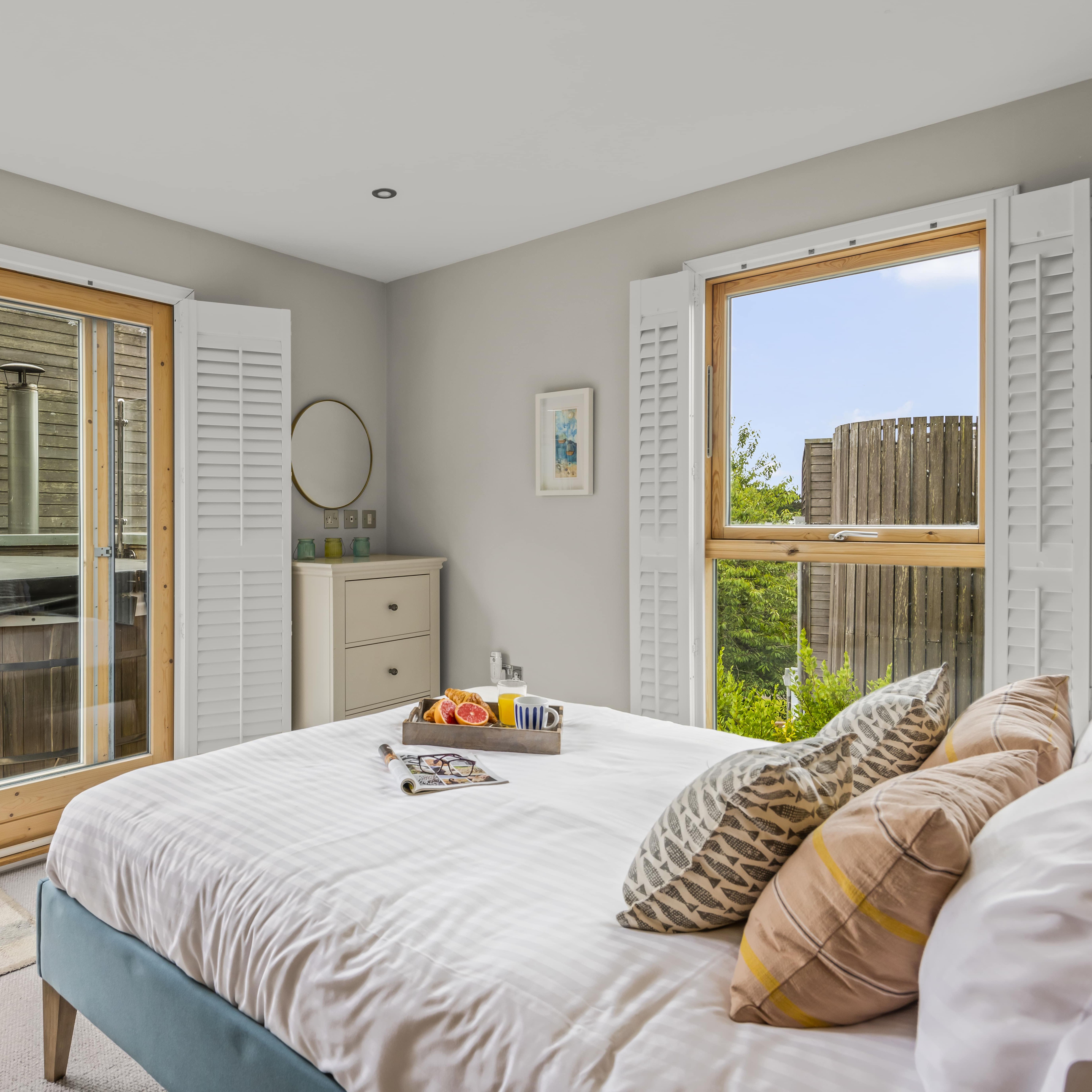 Bright bedroom with white bedding, wooden doors leading to outdoor wooden hot tub, and window shutters.