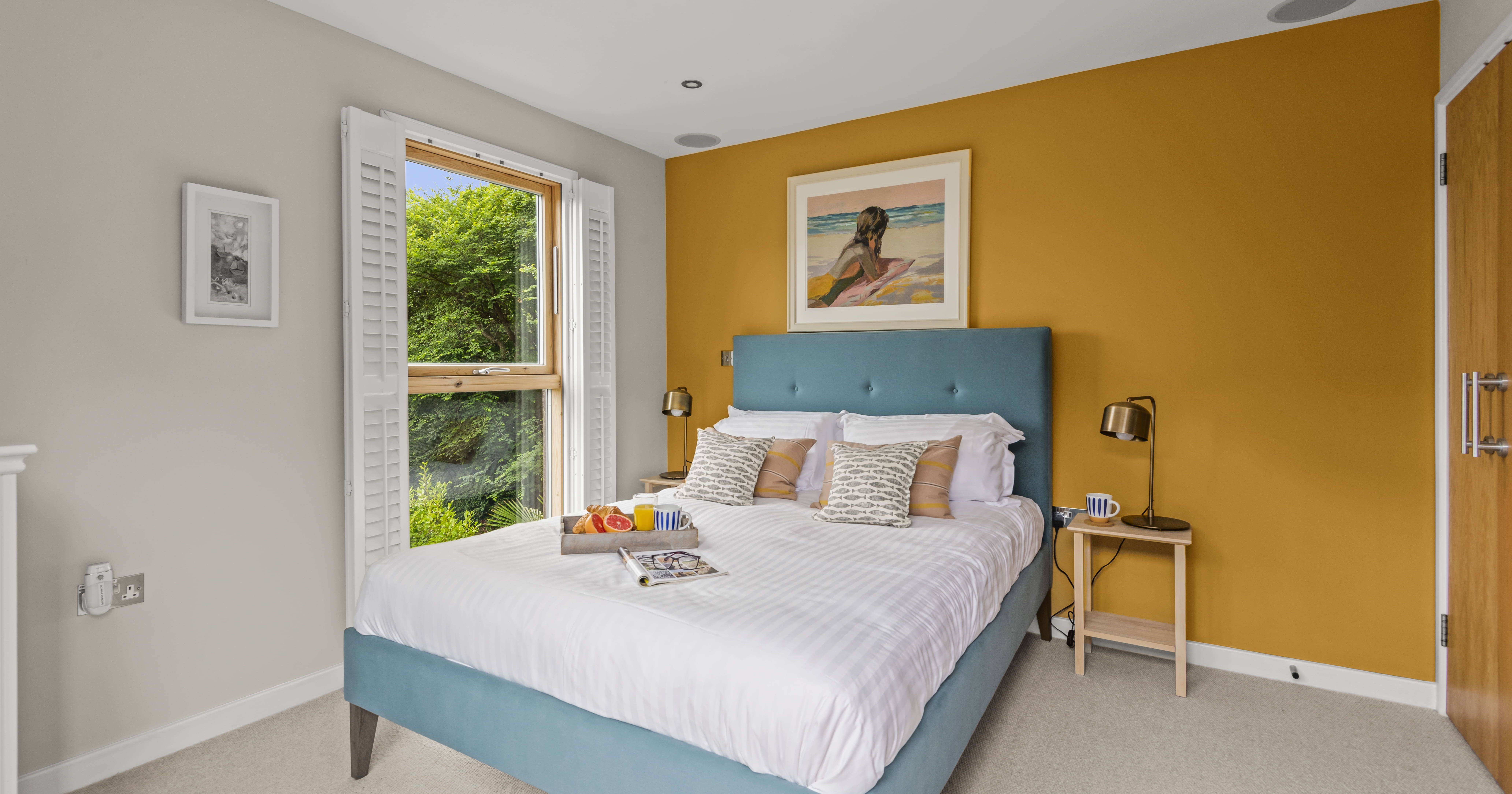 Modern bedroom with teal bed, mustard yellow accent wall, and window with white shutters