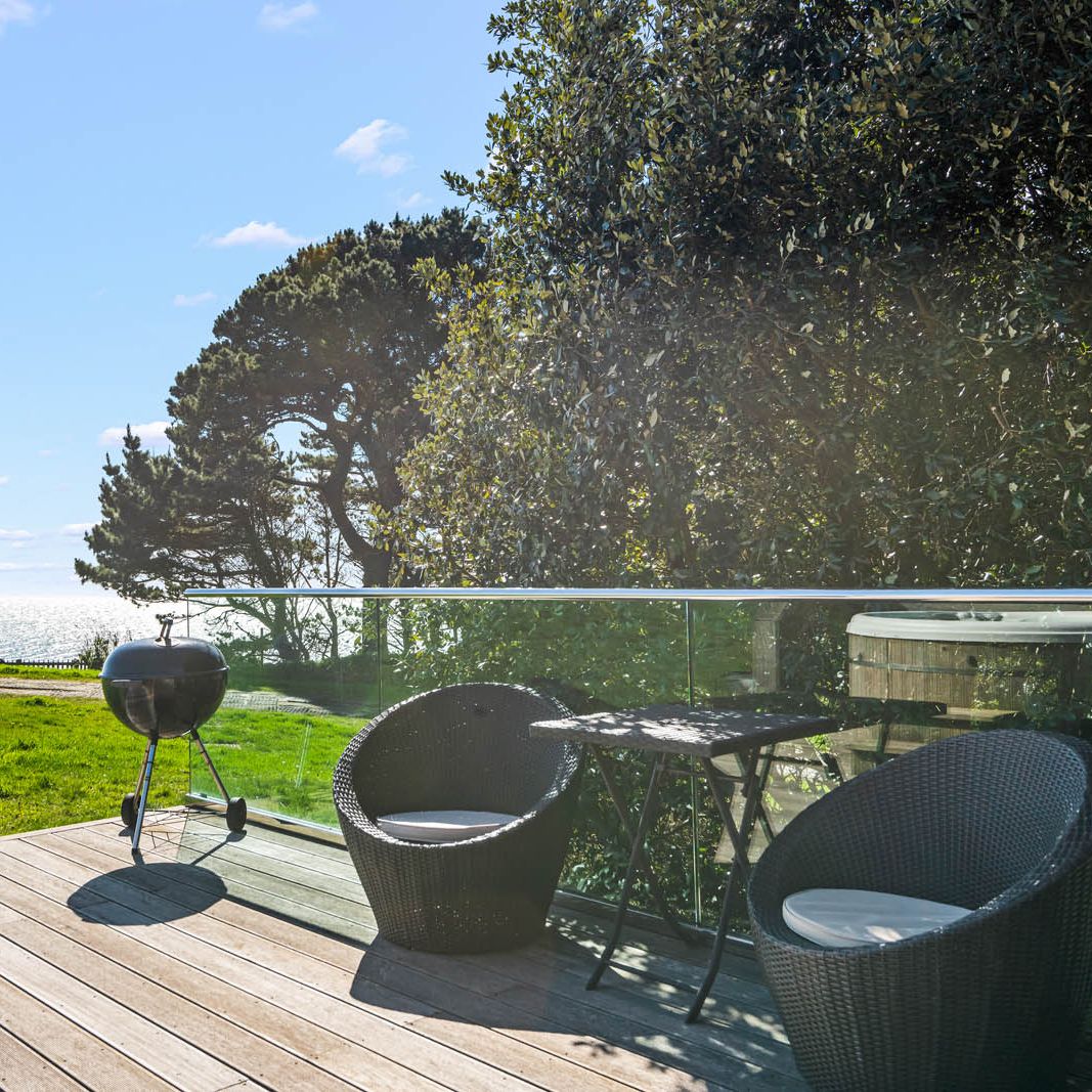 Sunlit wooden deck with two wicker chairs, a small table and a barbecue overlooking the sea