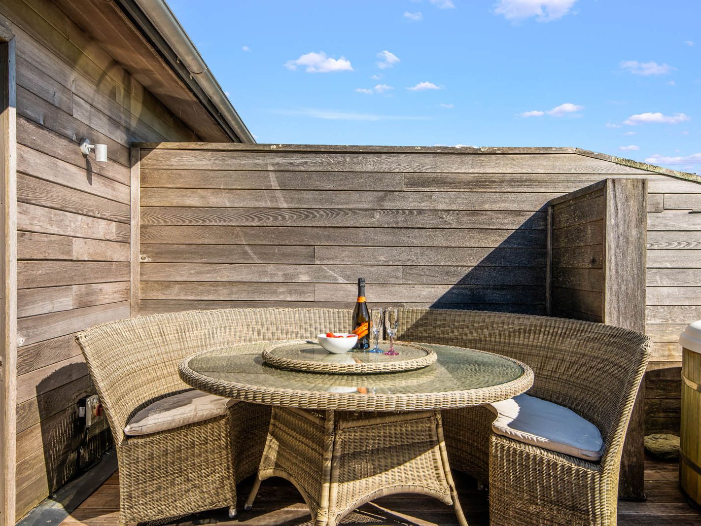 Wicker patio seating with round glass-topped table set with wine and glasses on a wooden deck