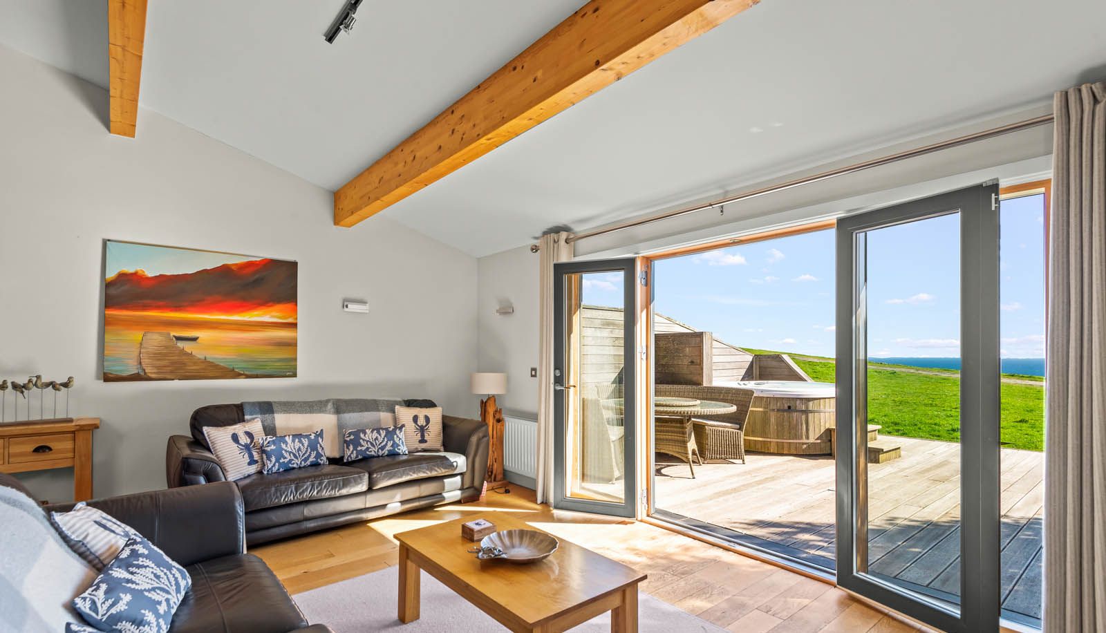 Bright living room with leather sofas, wooden beams and open sliding doors to a deck with a hot tub and sea view