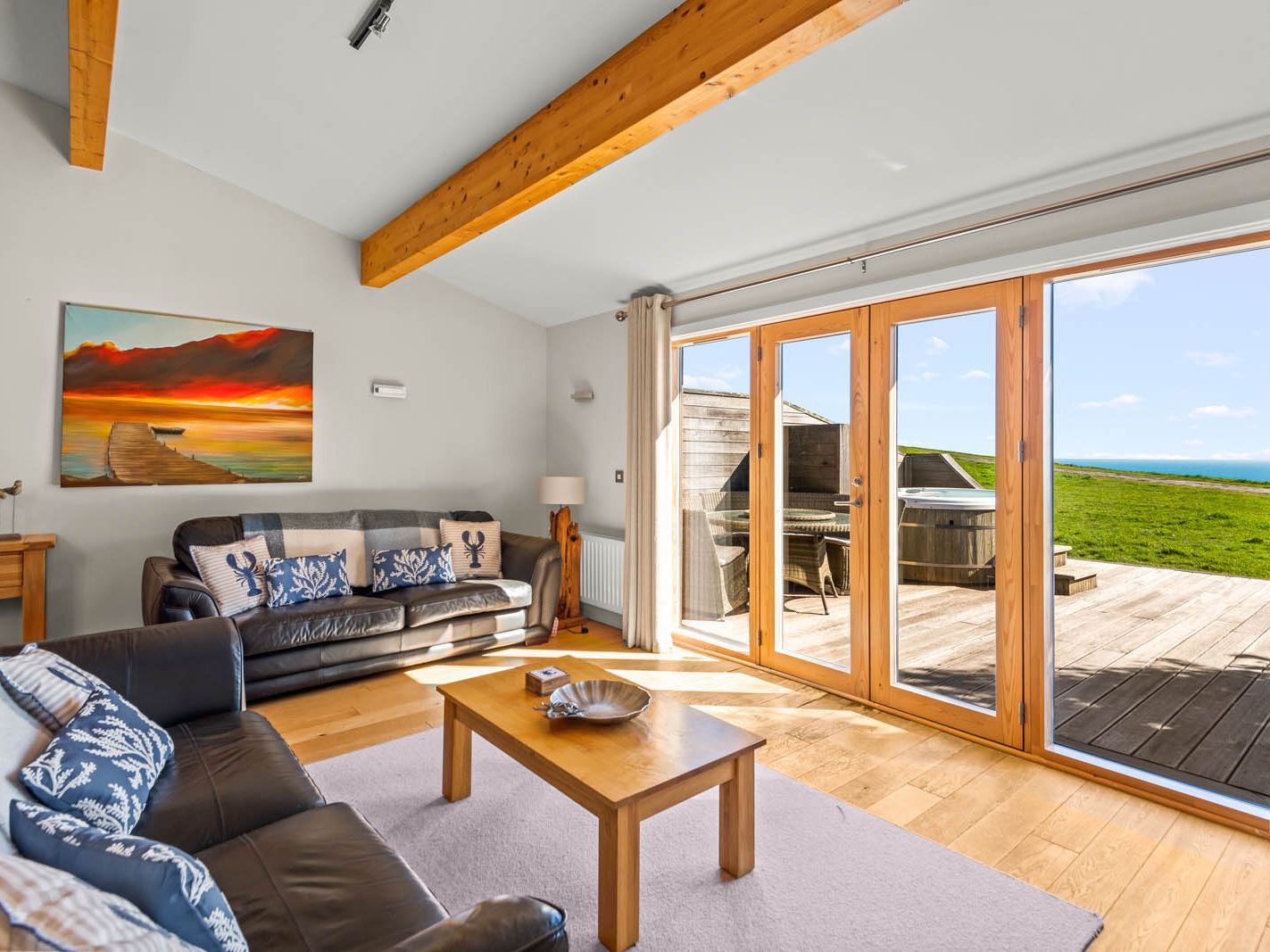 Bright coastal living room with leather sofas and wooden beams, opening through large glass doors onto a deck with sea view