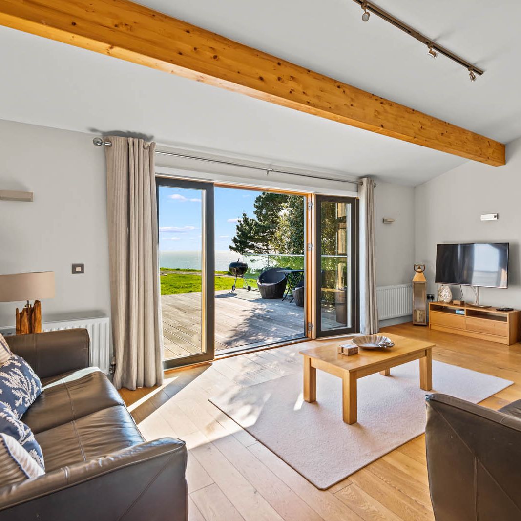 Bright living room with exposed wooden beams, leather sofas and sliding doors opening onto a deck with sea views