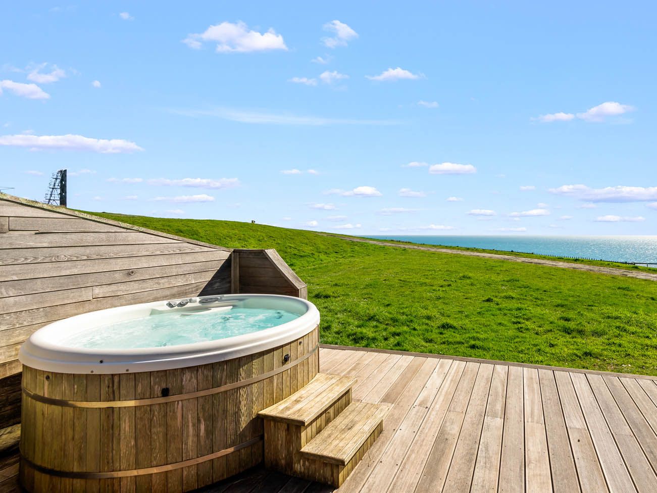 Wooden hot tub on a deck overlooking a grassy slope and the sea under a blue sky