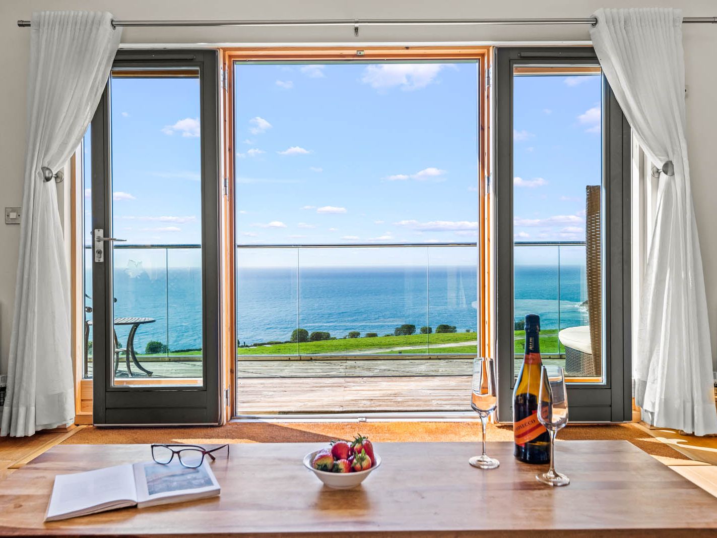 Open French doors with white curtains overlooking the sea and balcony, with champagne, glasses and strawberries on a table