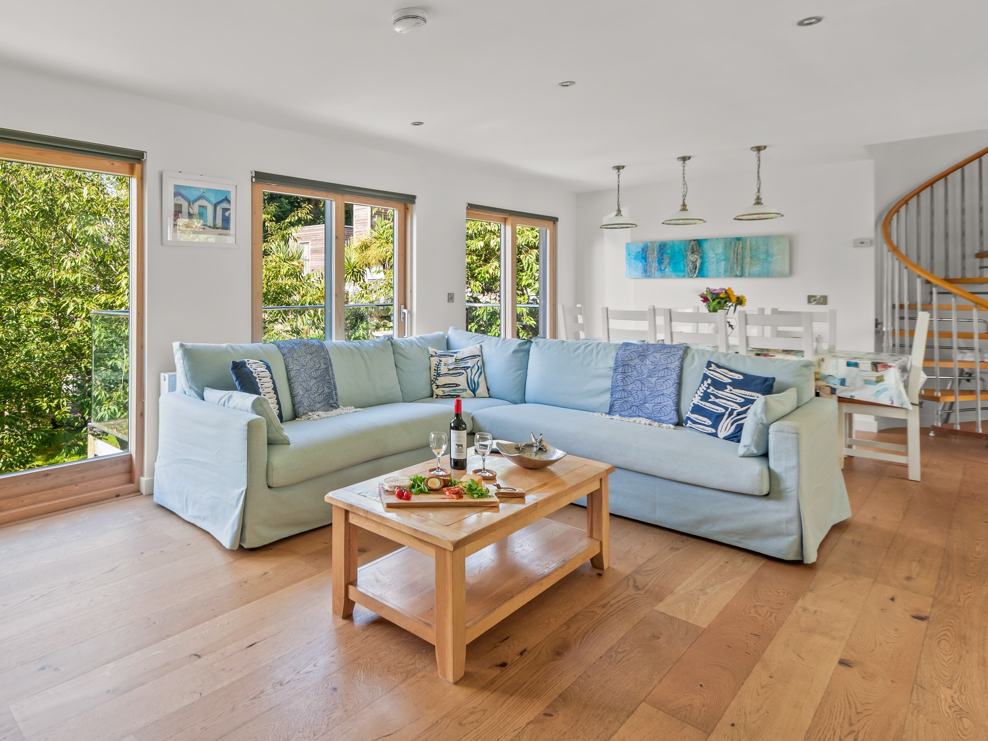 Bright open-plan living room with pale blue corner sofa, wooden coffee table and large windows overlooking trees