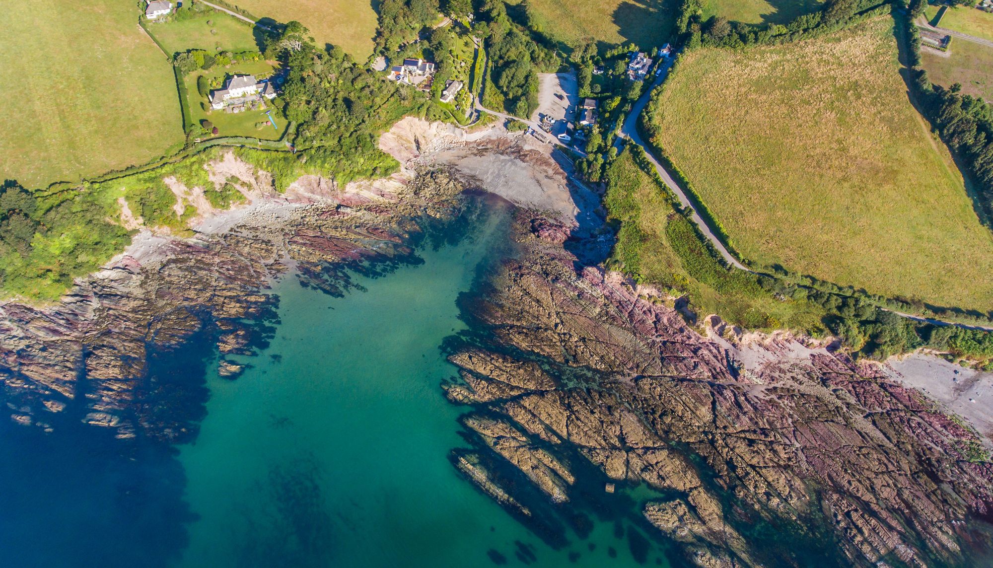 Aerial view of a rocky coastline and small cove with turquoise sea beside green fields and houses