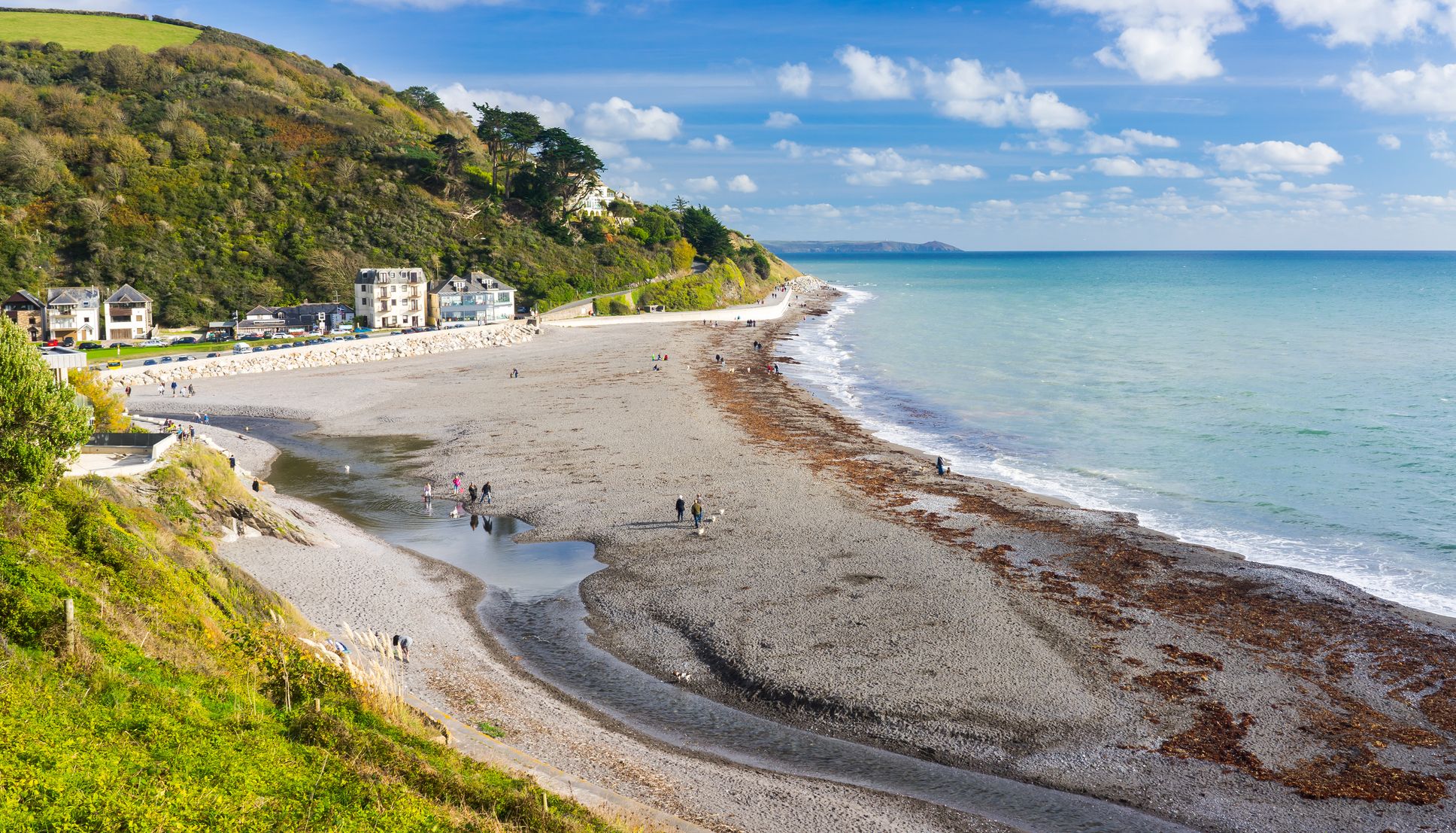 Coastal view of a long pebble beach beside green cliffs and seaside buildings under a blue sky