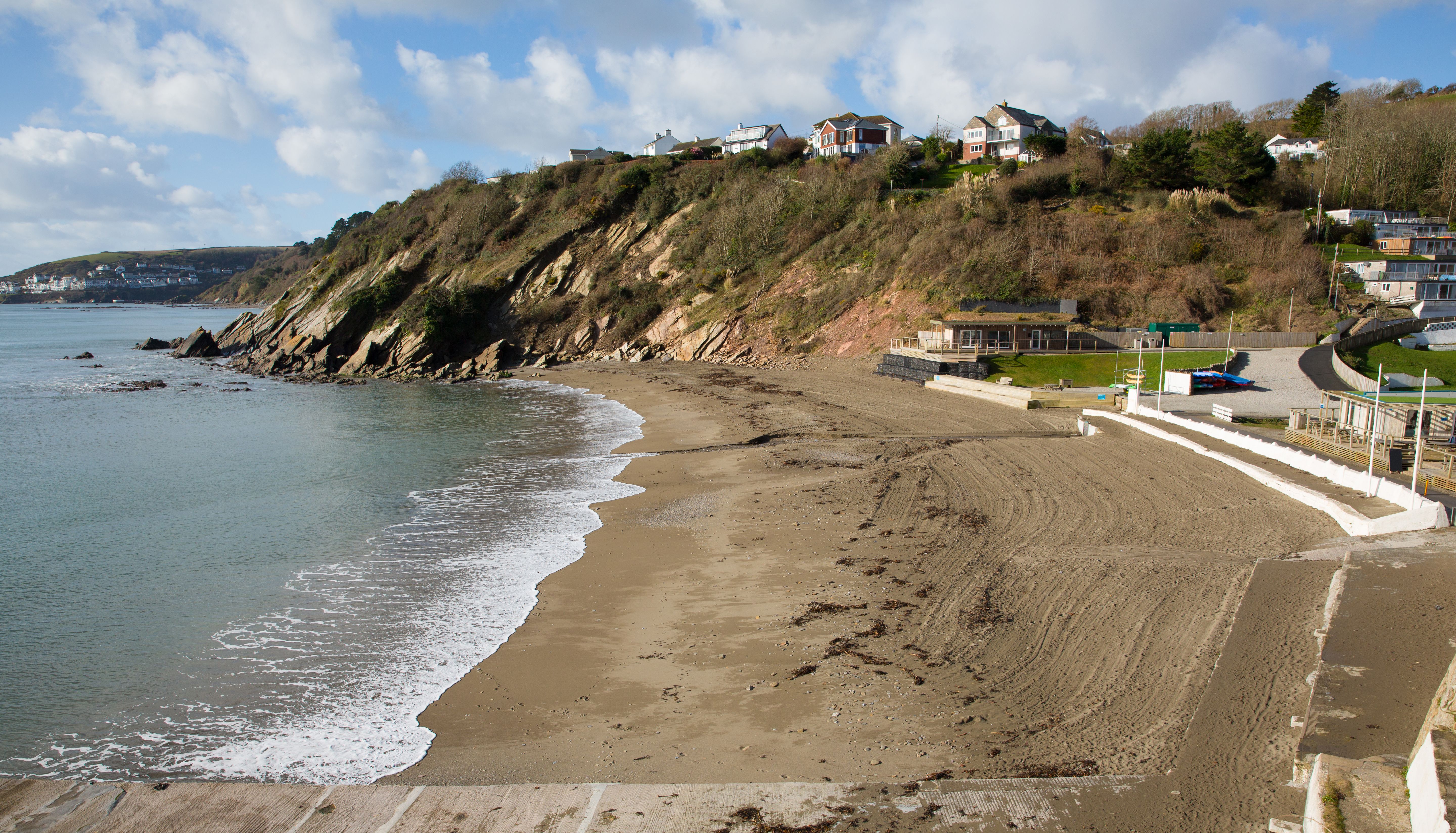 Curving sandy beach below a rocky cliff with seaside houses under a partly cloudy sky