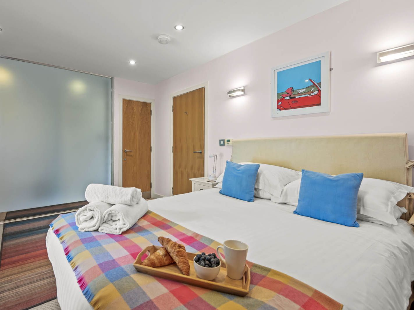 Bright modern bedroom with double bed, blue cushions and breakfast tray, next to an en-suite behind a frosted glass partition