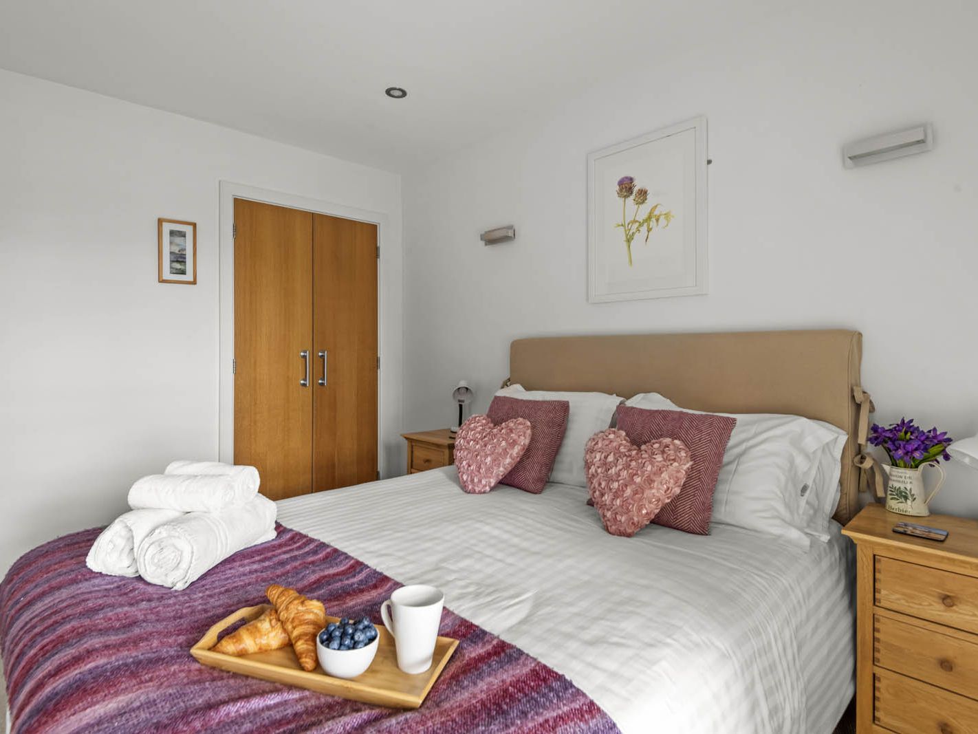 Bright bedroom with a double bed, heart-shaped cushions, folded towels and a breakfast tray with croissants and blueberries