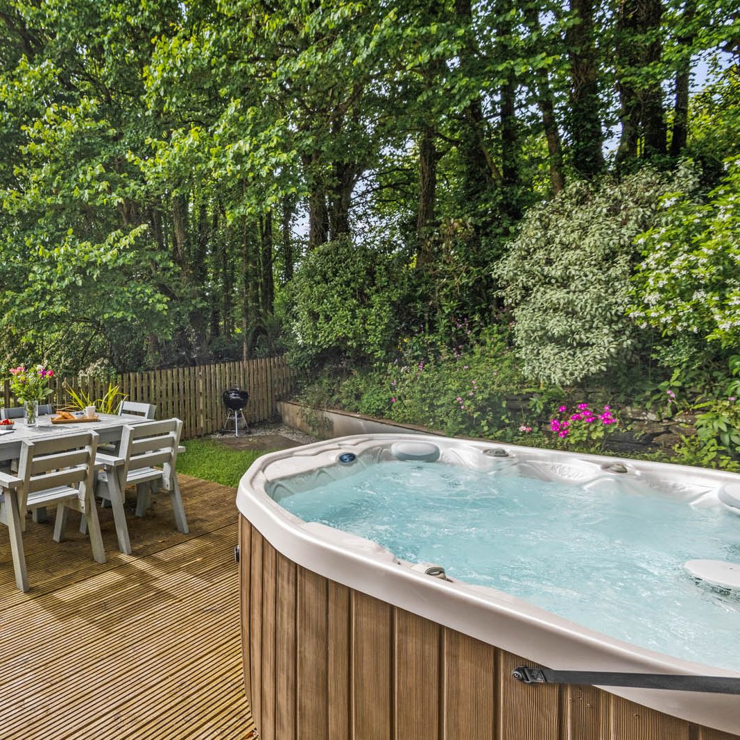 Outdoor wooden deck with dining table and chairs beside a hot tub, surrounded by trees and garden plants