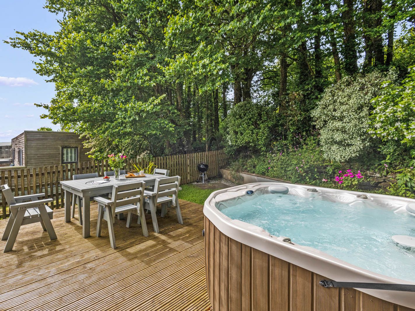 Outdoor wooden deck with dining table and chairs beside a hot tub, surrounded by trees and garden plants