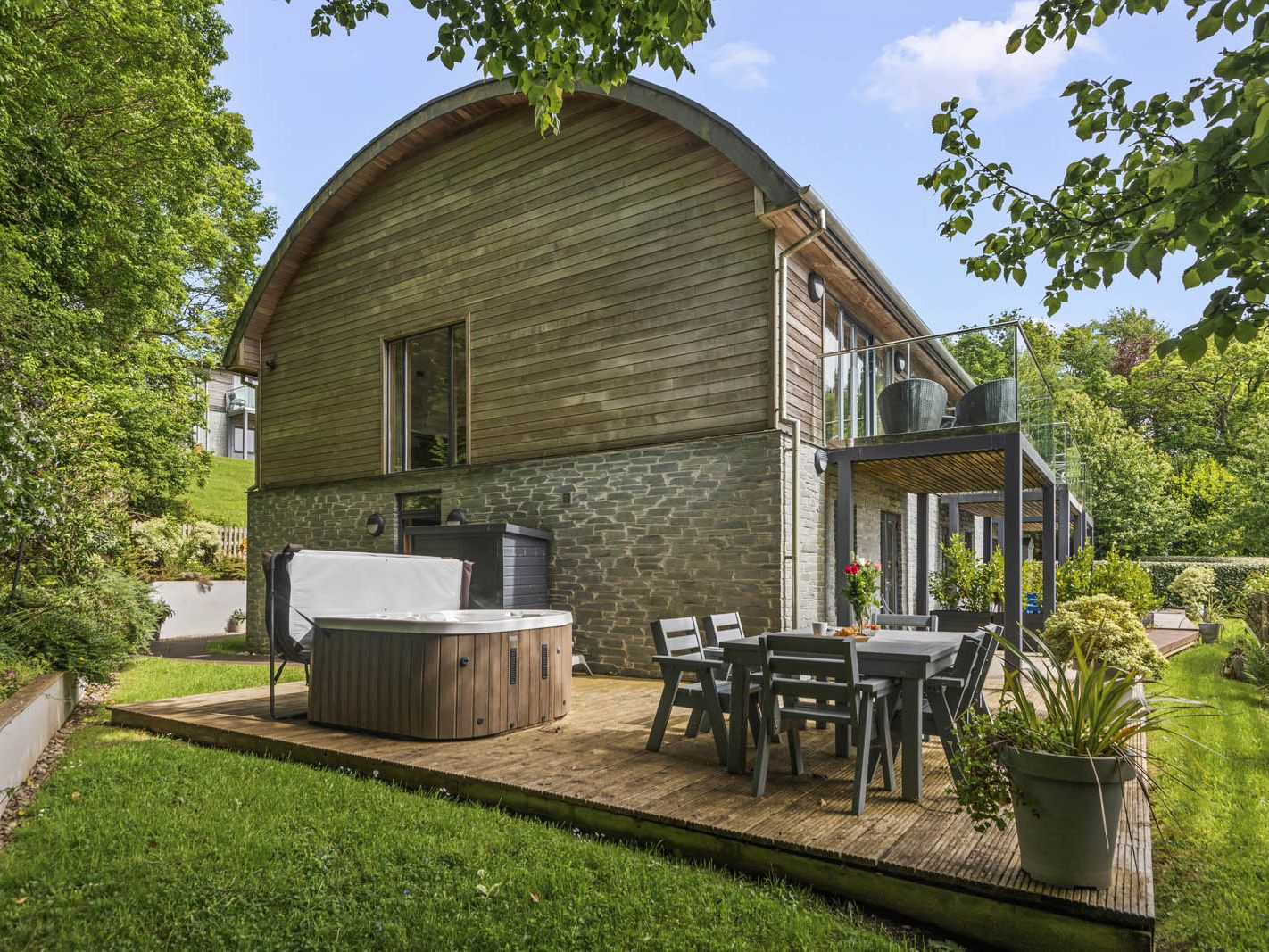 Modern timber-and-stone house with a curved roof, balcony and garden decking with hot tub and outdoor dining set