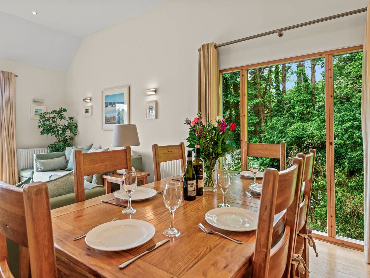 Wooden dining table set with plates and wine glasses beside large window overlooking green woodland