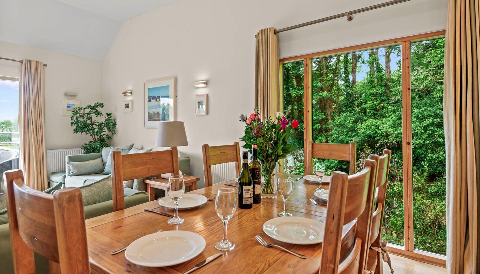 Wooden dining table set with plates and wine glasses beside large window overlooking green woodland