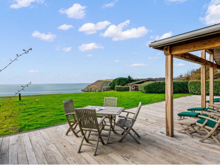 Wooden deck with outdoor table and chairs overlooking a green lawn and the sea under a blue sky