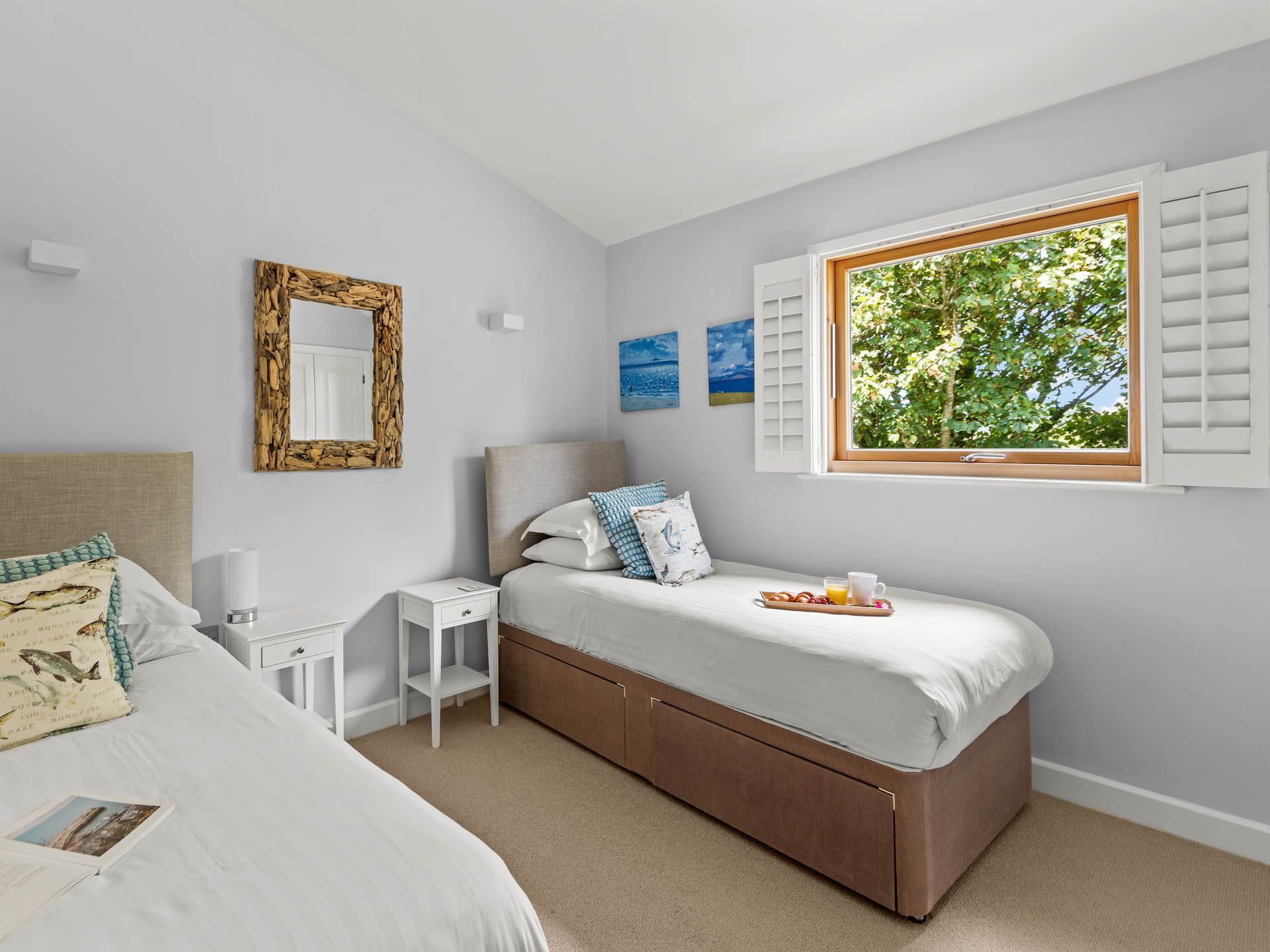 Bright twin bedroom with two single beds, white walls, bedside tables and a window with shutters overlooking green trees