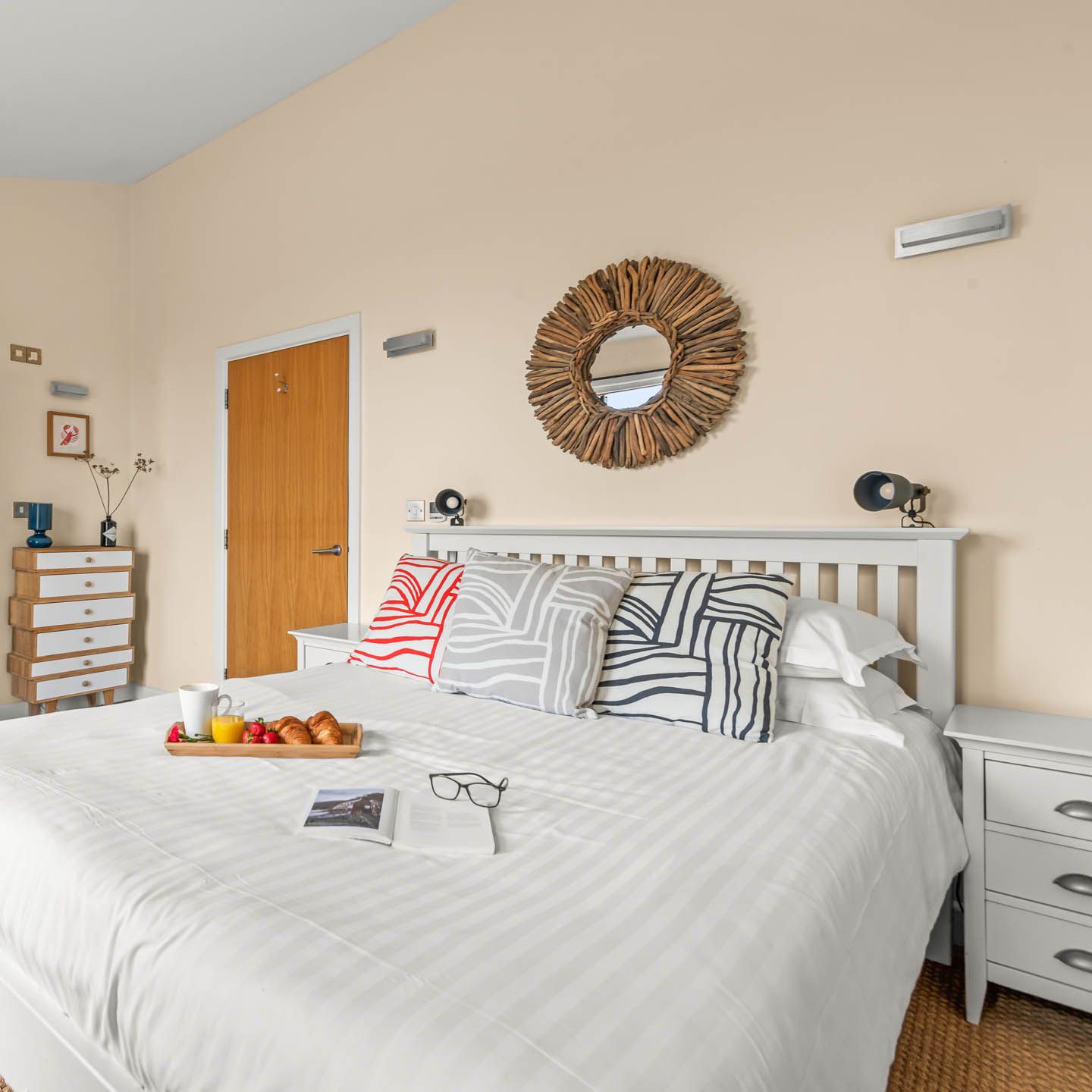 Bright bedroom with white bed, patterned cushions and driftwood-framed mirror, with open door to en-suite shower